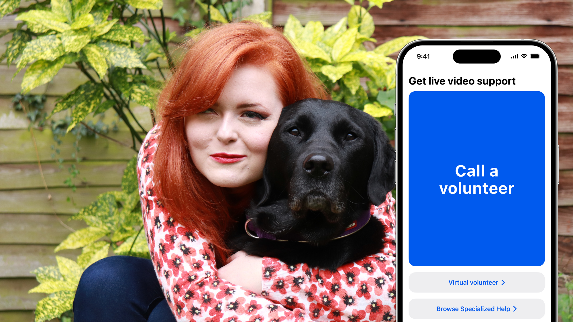 Lucy is crouching down and hugging a black Labrador in front of a garden wall. Next to her and the dog is a large iPhone screen showing the homepage of the Be My Eyes app, which is photoshopped into the picture.