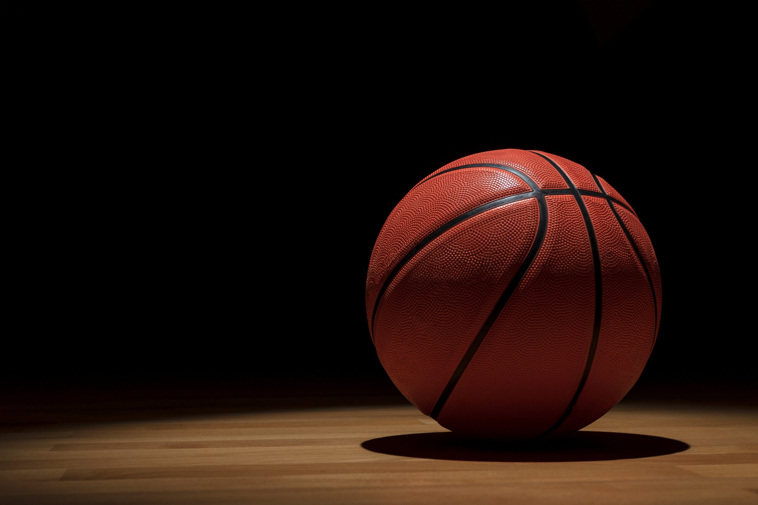ISOLATED BASKETBALL BALL ON WOODEN COURT WITH SPOTLIGHTS FROM ABOVE AND DARK BACKGROUND WHEN THE GAME IS OVER.