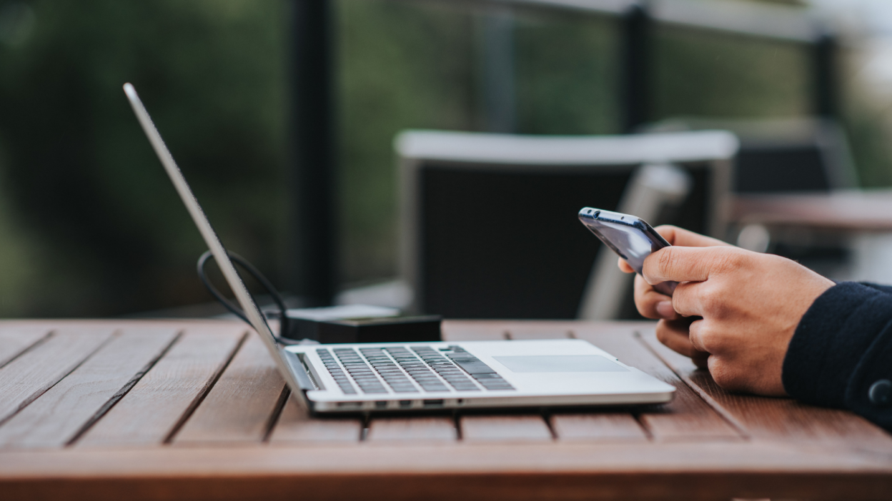 person using phone and laptop outside at picnic table