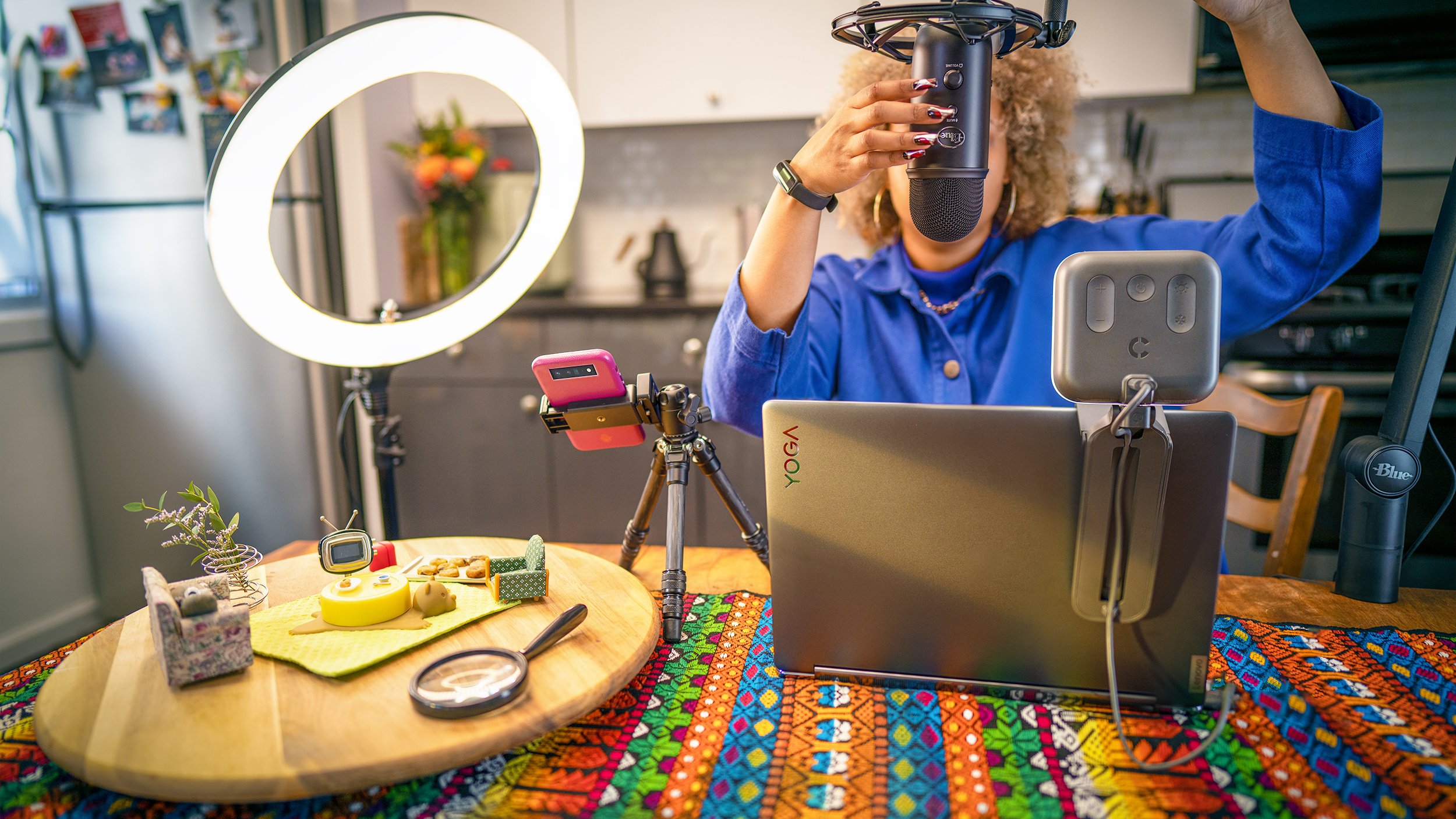 Videographer sitting at a table for a videoshoot makes adjustments to the microphone; a ringlight, laptop, and smartphone are set up on the table, along with a tiny kitchen set, featuring a miniature couch, TV, sheet of cookies, and table. 