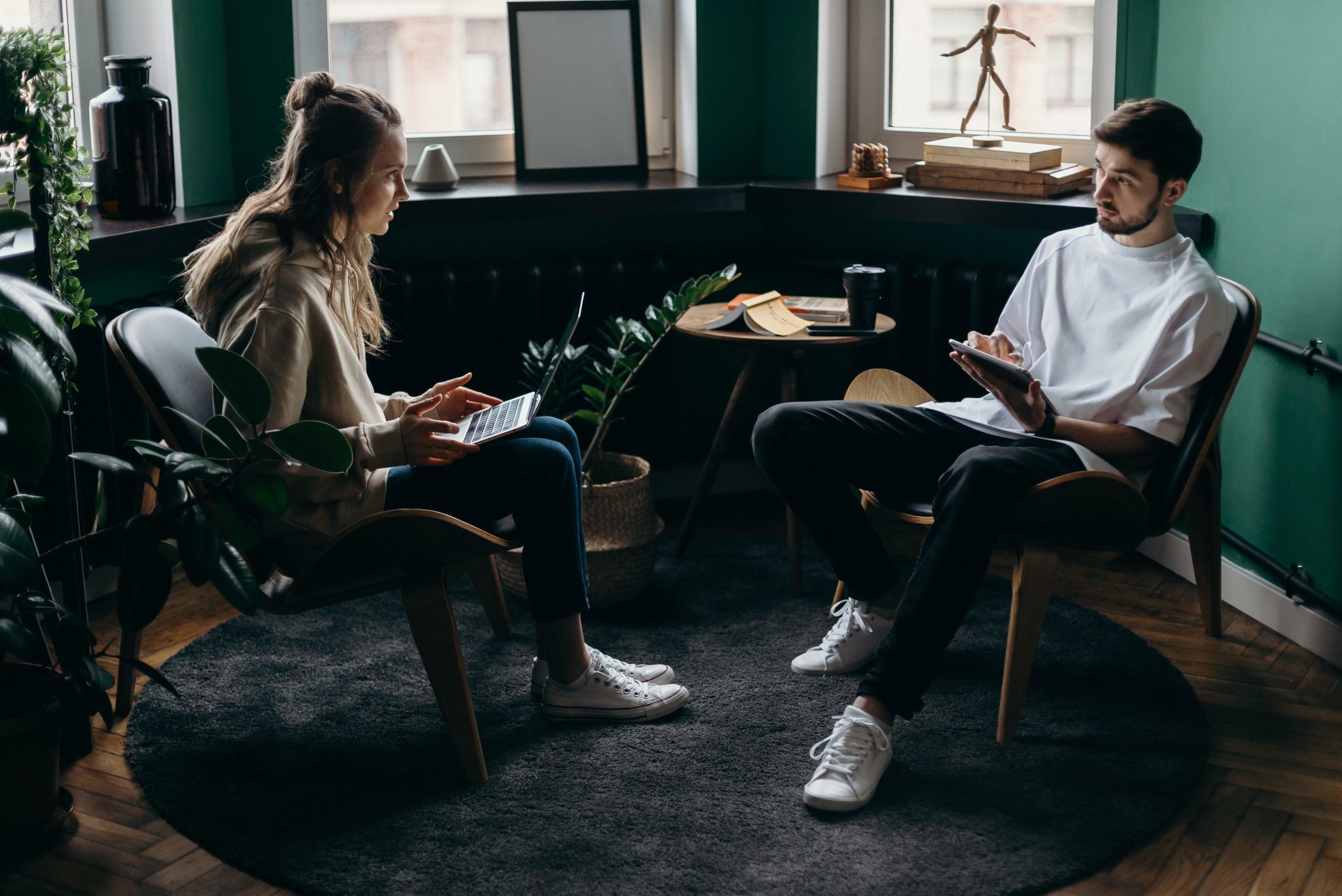 two people are sitting in a corner of a darkly lit room by bay windows, each holding a laptop and iPad air respectively