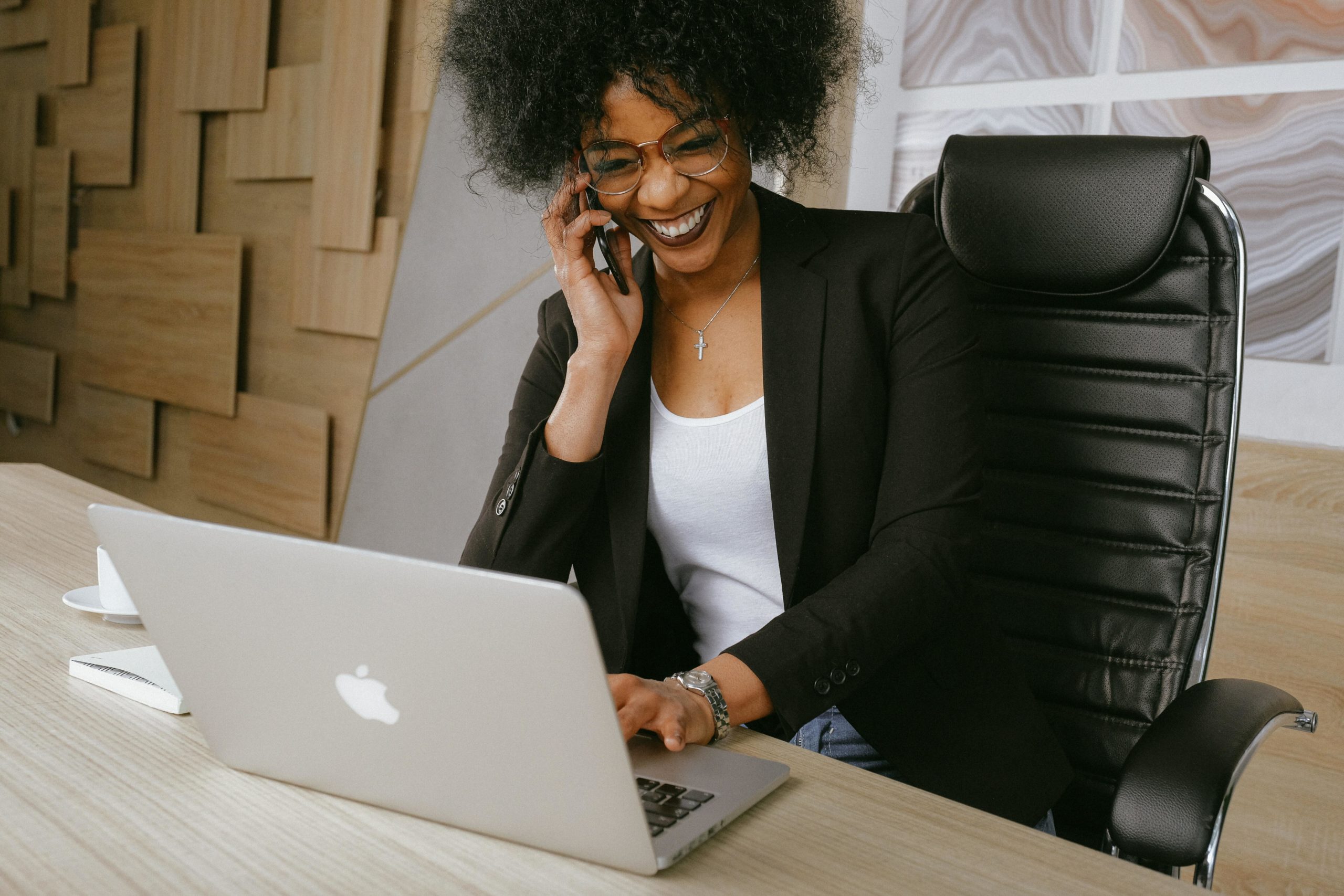 Business woman smiles on an iPhone while typing on a Mac laptop at a desk.