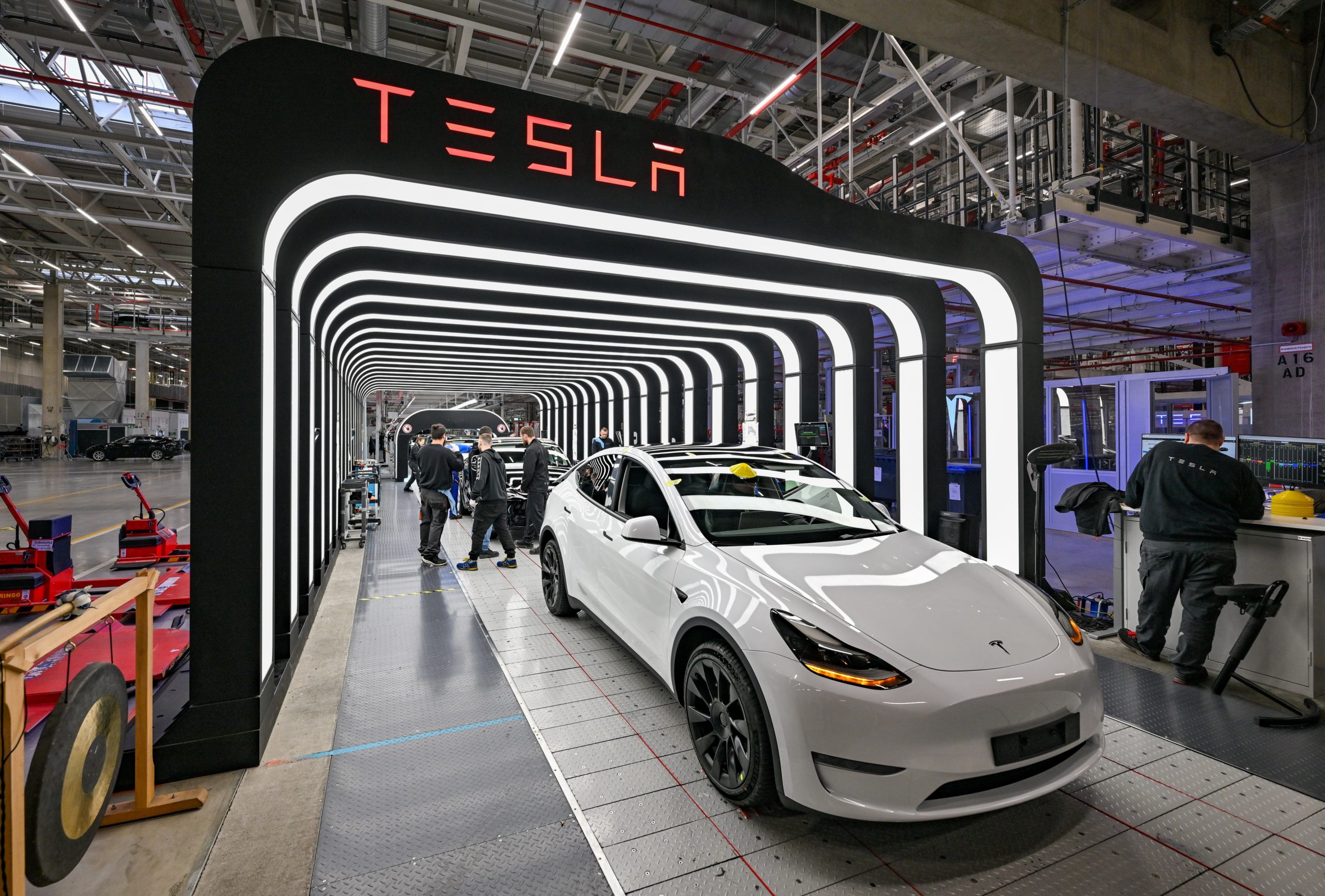 A white Tesla Model Y is parked in the glowing archway of a Tesla inspection bay. Employees work behind the vehicle.