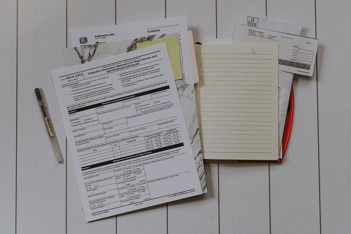 tax documents, forms, manila envelopes, ruled paper, and receipts are laid out against white-painted wood paneling. one muji ballpoint pen is lying beside the forms and documents