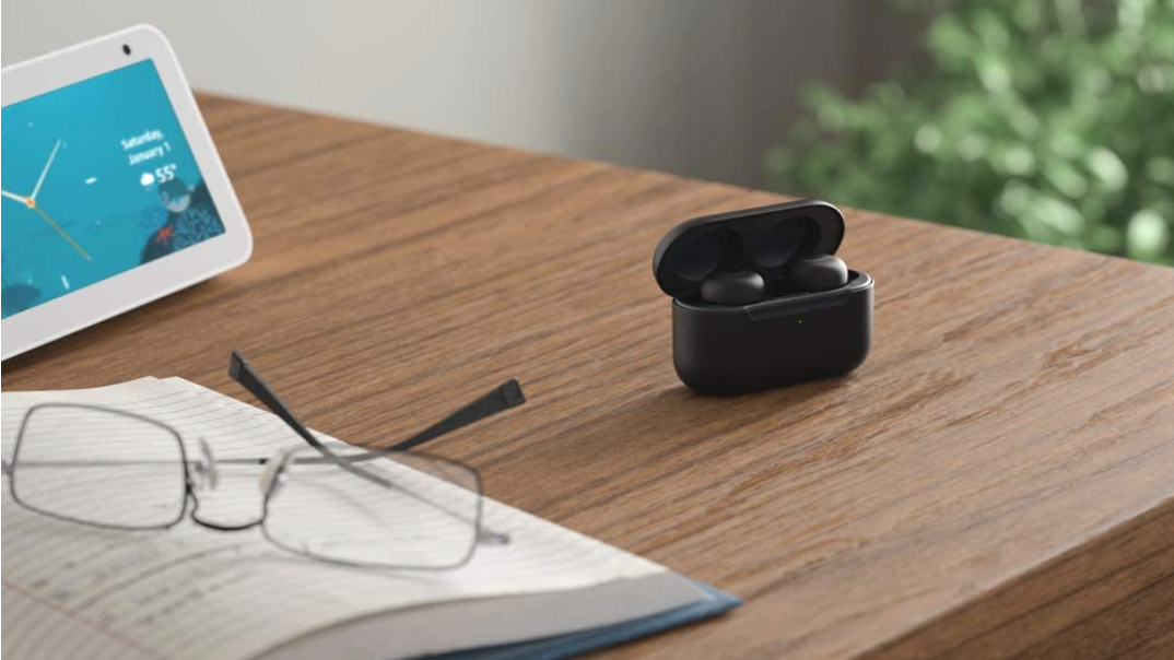 Black Amazon Echo Buds on a brown wood desk with a pair of glasses on top of a book on bottom left, tablet on upper left side