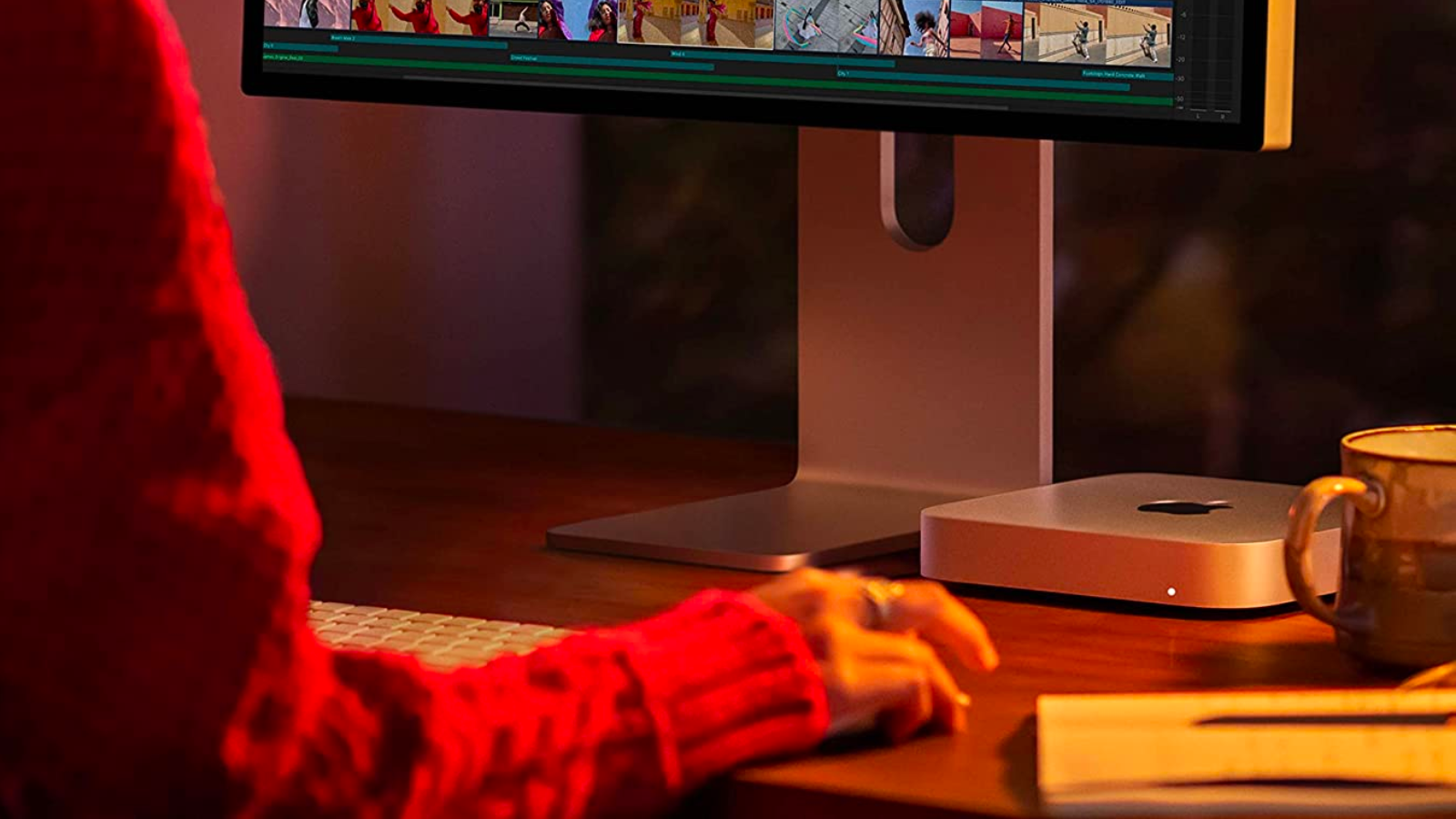 a close-up of a mac mini underneath an apple studio display next to a mug, a notebook, and a woman's hand on a mouse in dim, moody lighting