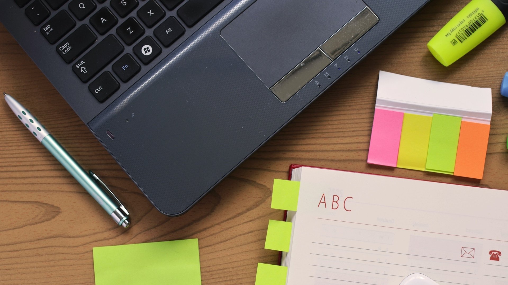 A laptop, a pen, a highlighter, and notebook rest on a desk.