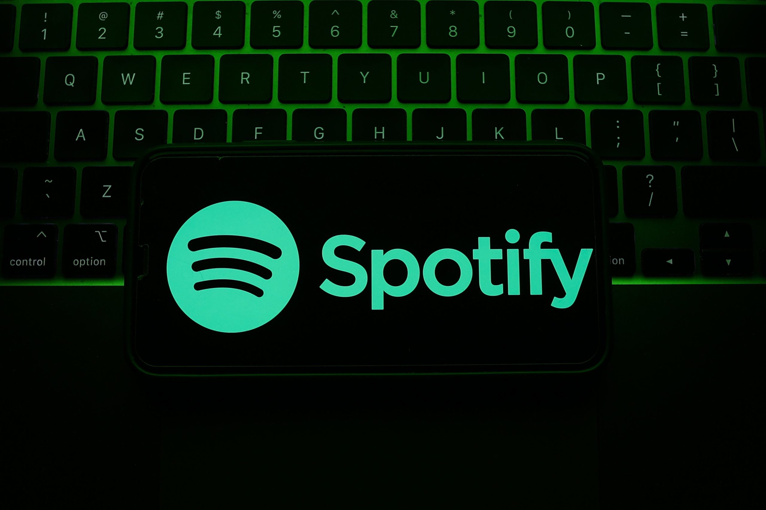 A phone with Spotify open overlaid over a laptop keyboard bathed in green light.