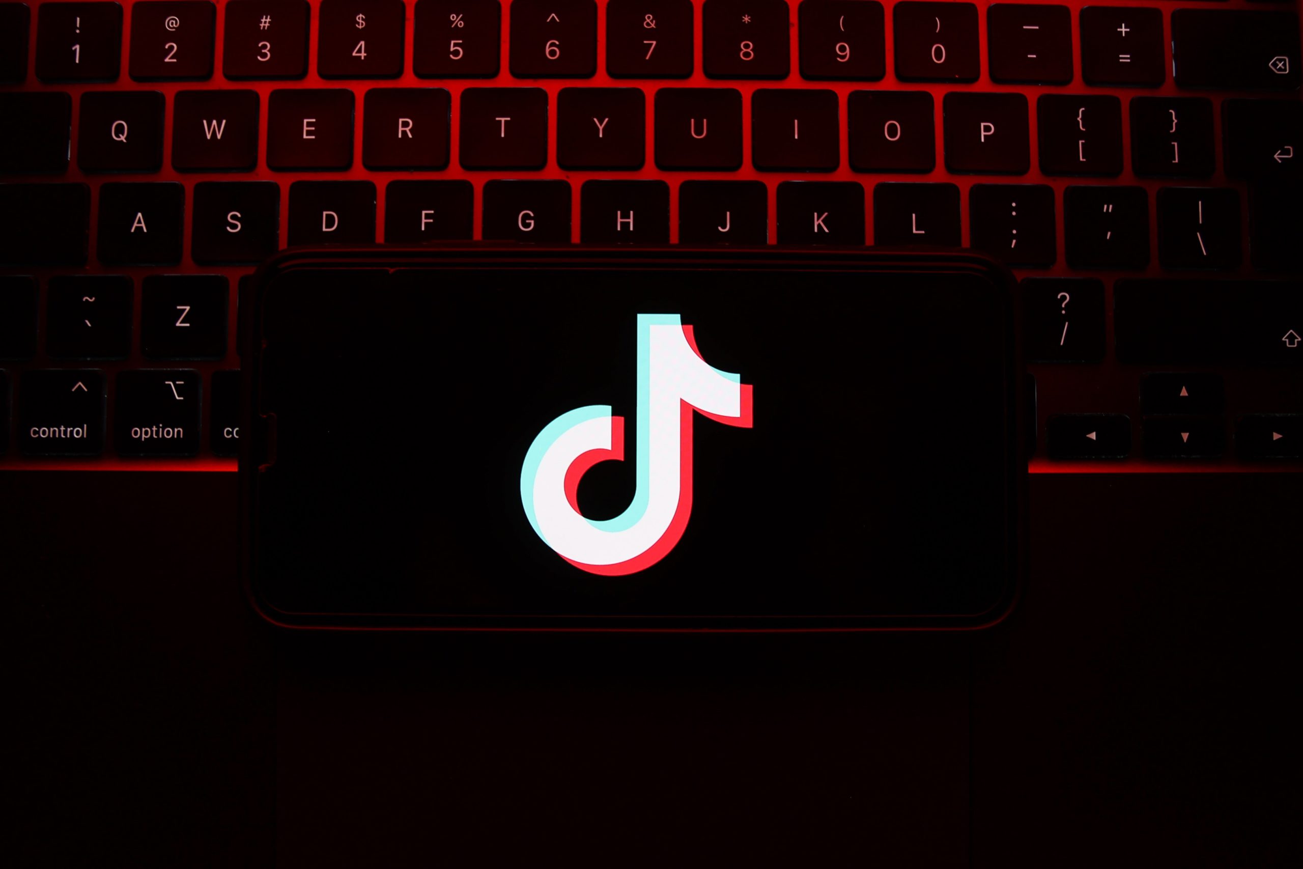 An iPhone screen showing the TikTok logo sitting on a laptop keyboard bathed in pink light. 