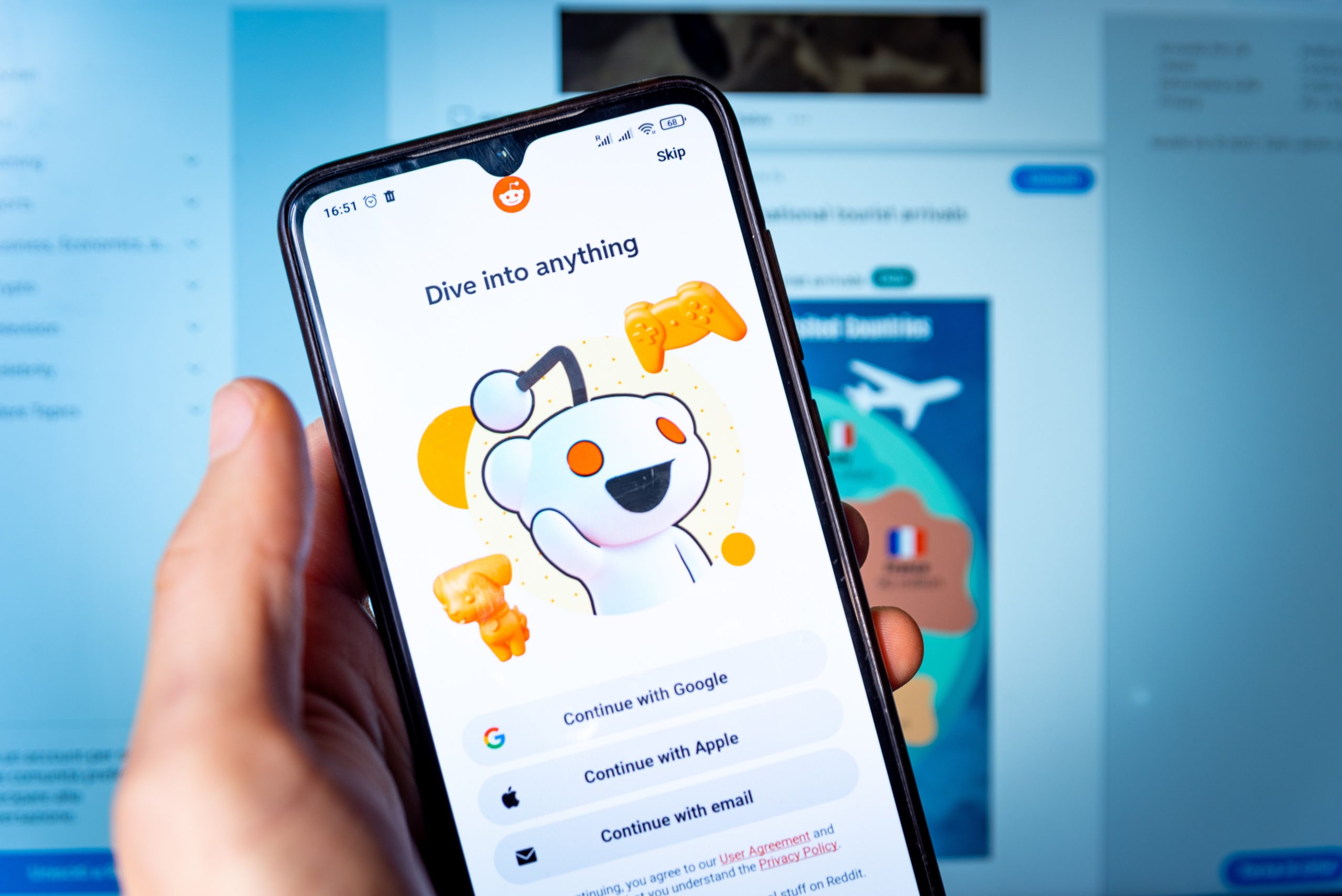 Reddit welcome page depicted on a smartphone being held by a hand in front of a computer monitor