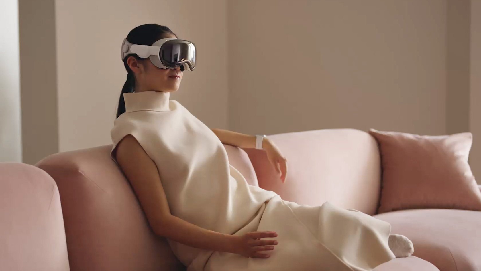 An Asian woman sits on a light pink couch, She is in a white dress and is wearing the Vision pro on her head.