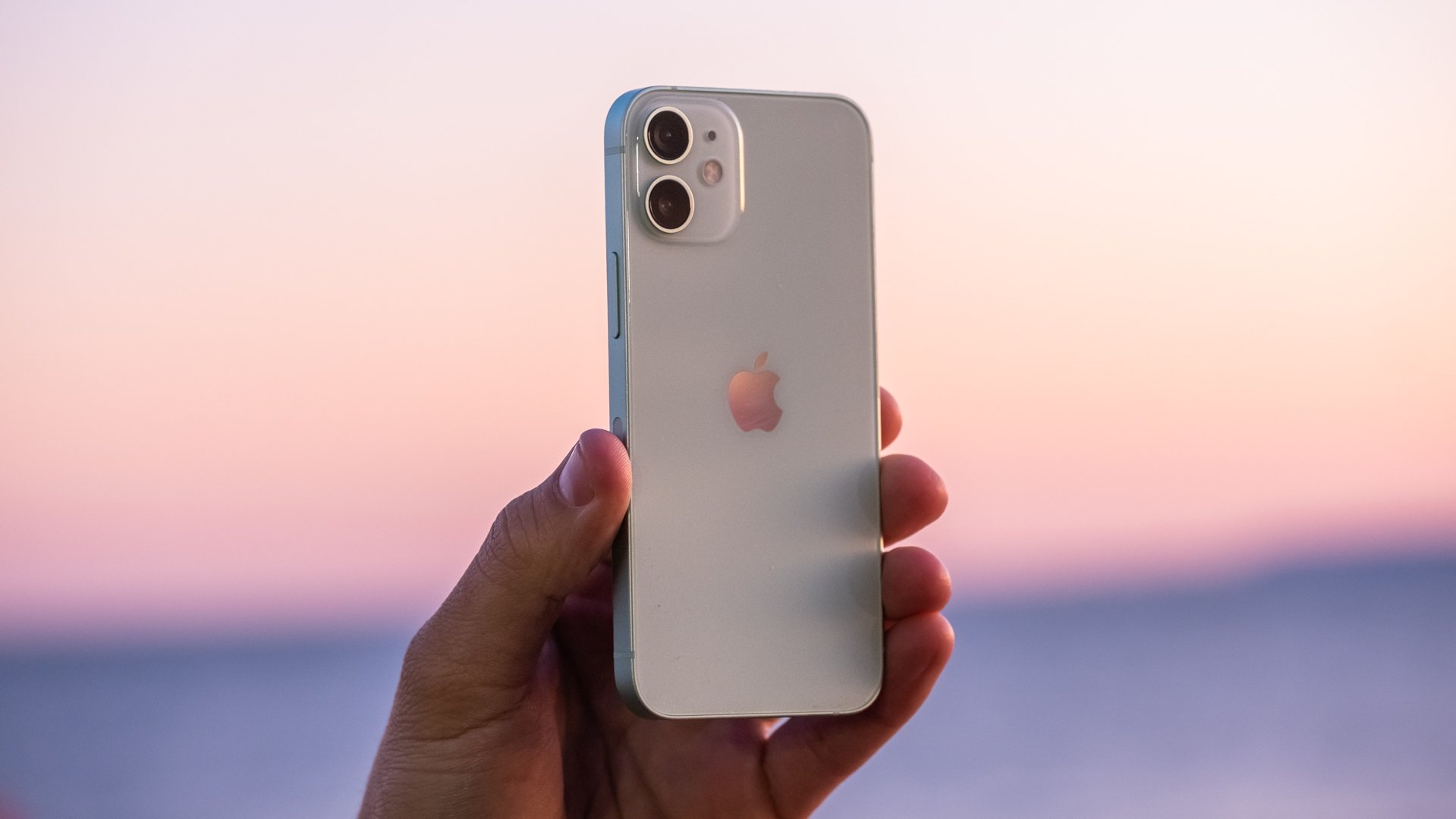 The Apple iPhone 12 Mini held up in someone's hand in front of a blurry outdoor background