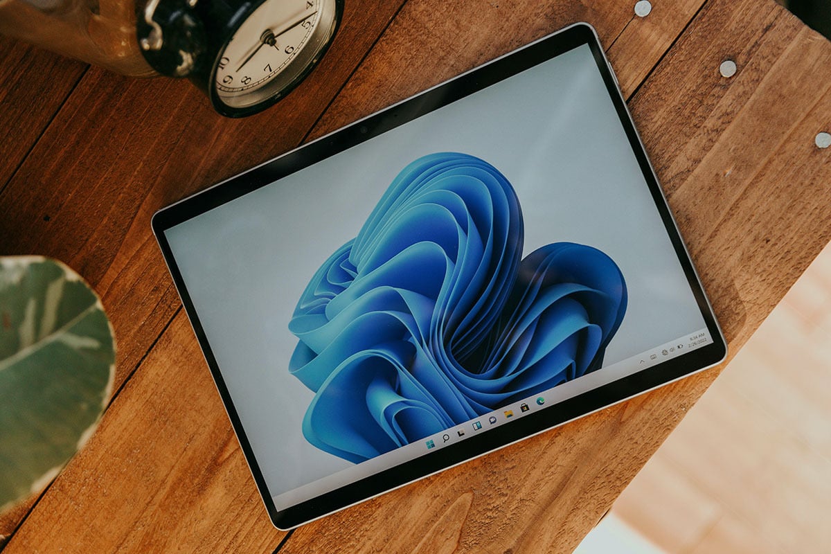 A tablet resting on a wooden desk surface surrounded by a clock and plant