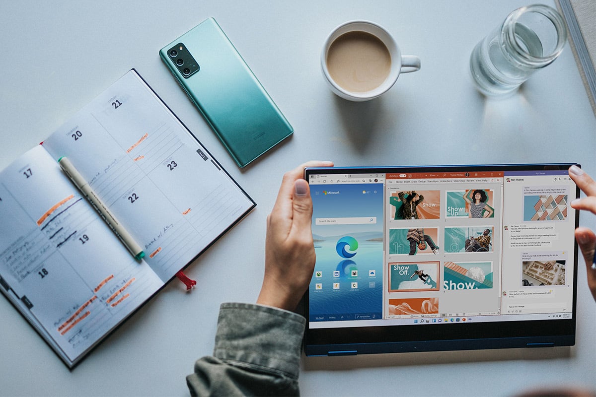 Someone's hands holding a tablet surrounded by a planner, coffee, phone, and glass of water