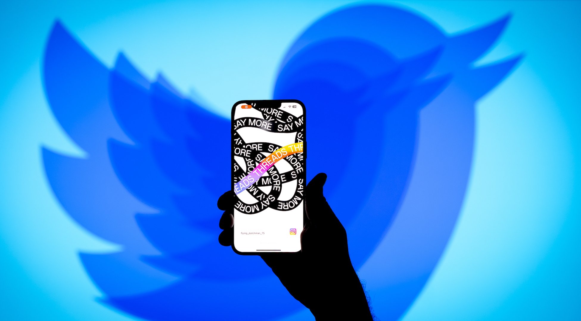 A shrinking Twitter logo behind a smartphone featuring the Threads app.