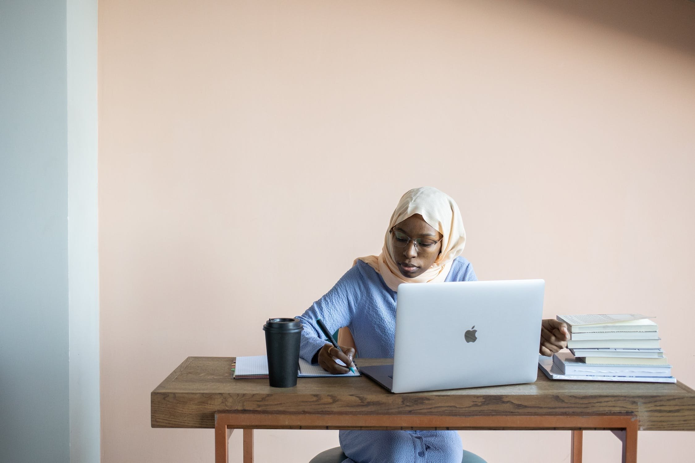 Woman sitting in front of an open laptop and writing notes with books and a cup of coffee nearby.