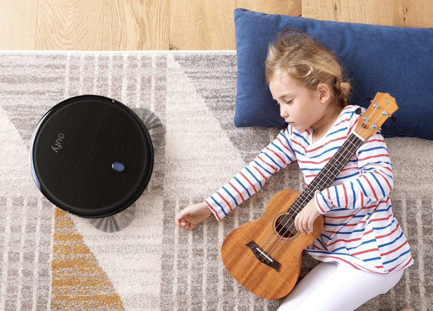 The Eufy BoostIQ RoboVac 11S on a carpet while a little girl sleeps next to it, holding a small guitar.
