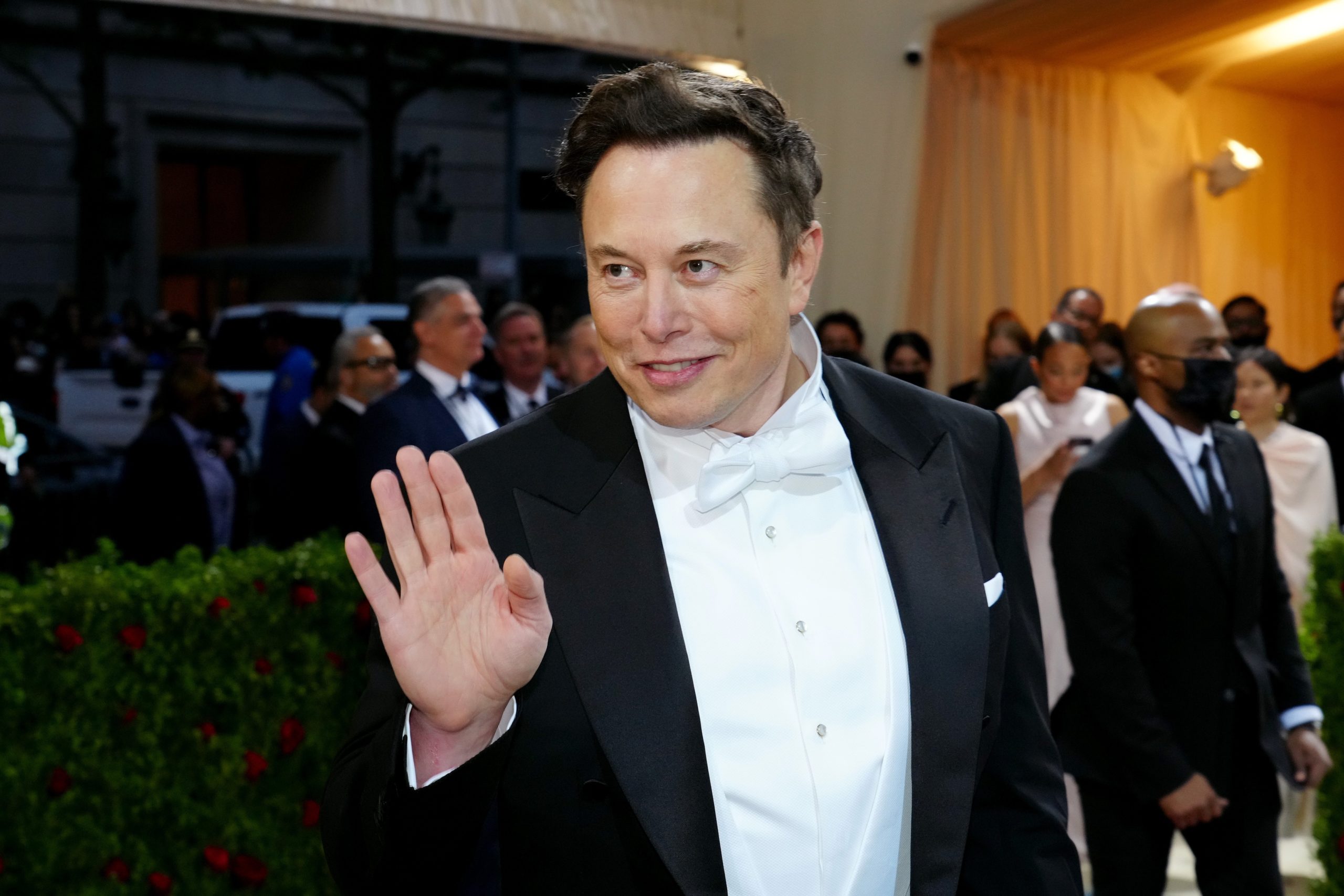 Elon Musk in a tuxedo at a formal event, slyly raising his hand.