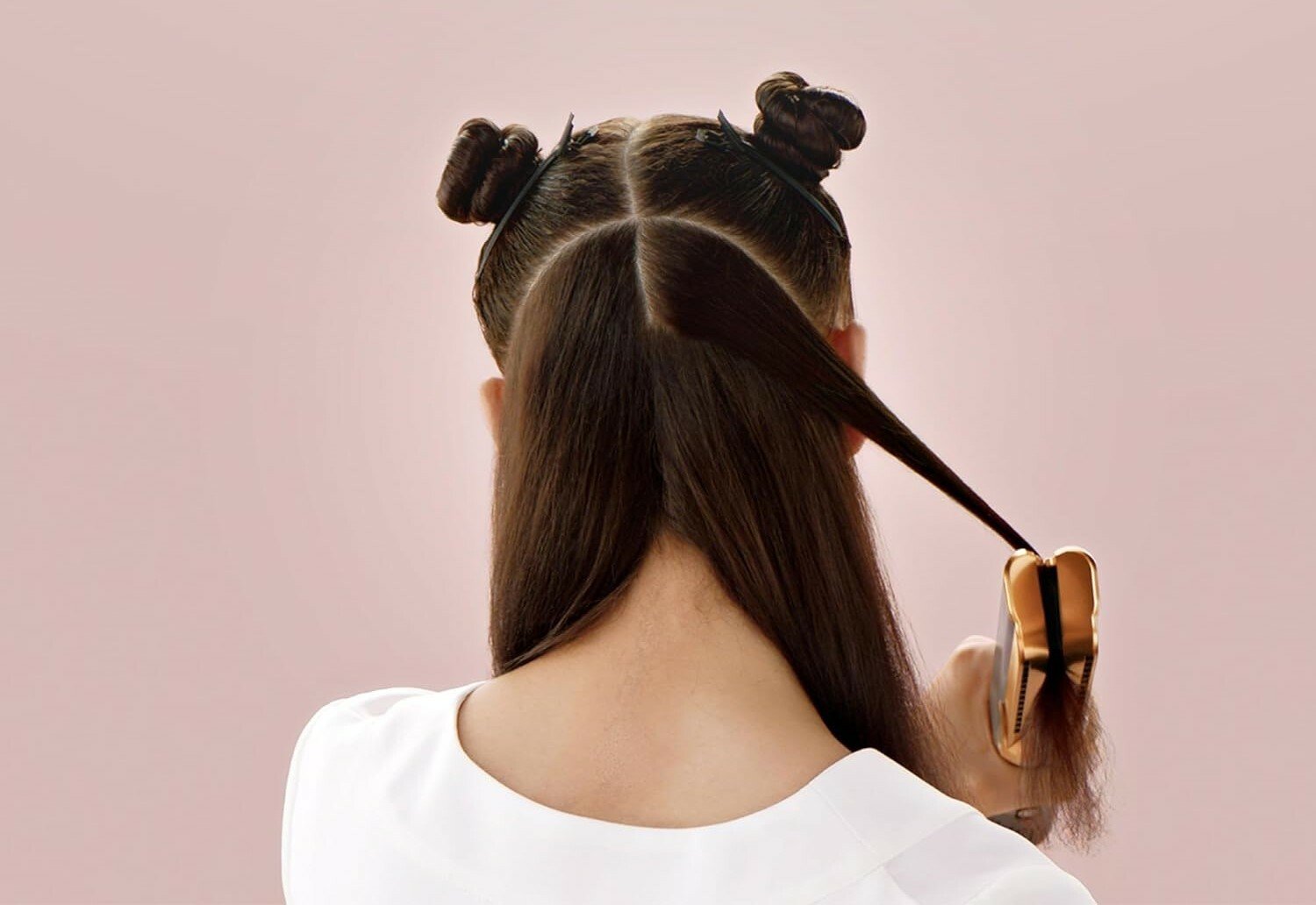 a person with long brown hair uses the dyson airstrait while standing in front of a pink background