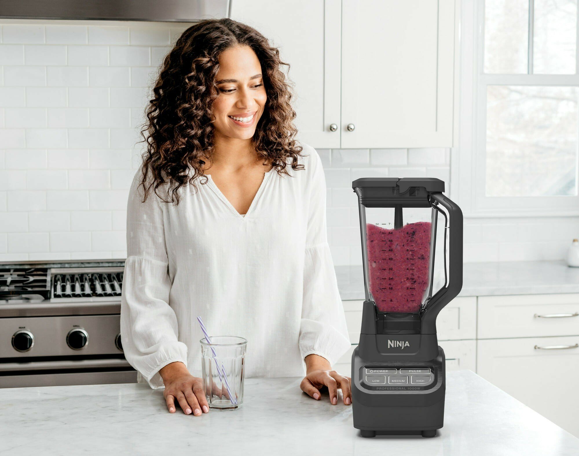 a woman stands behind a kitchen counter while looking at the ninja professional blender full of a purple colored smoothie in the blender's pitcher