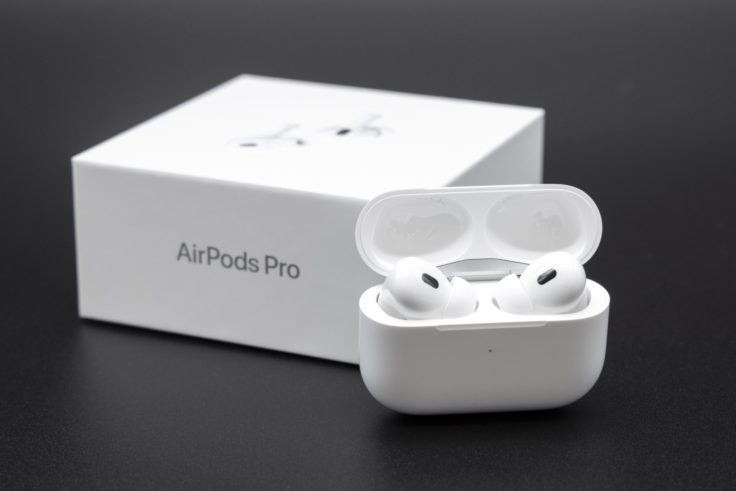 Product shot of Apple AirPods Pro 2nd generation inside the charging case next to the packaging box. Dark background.