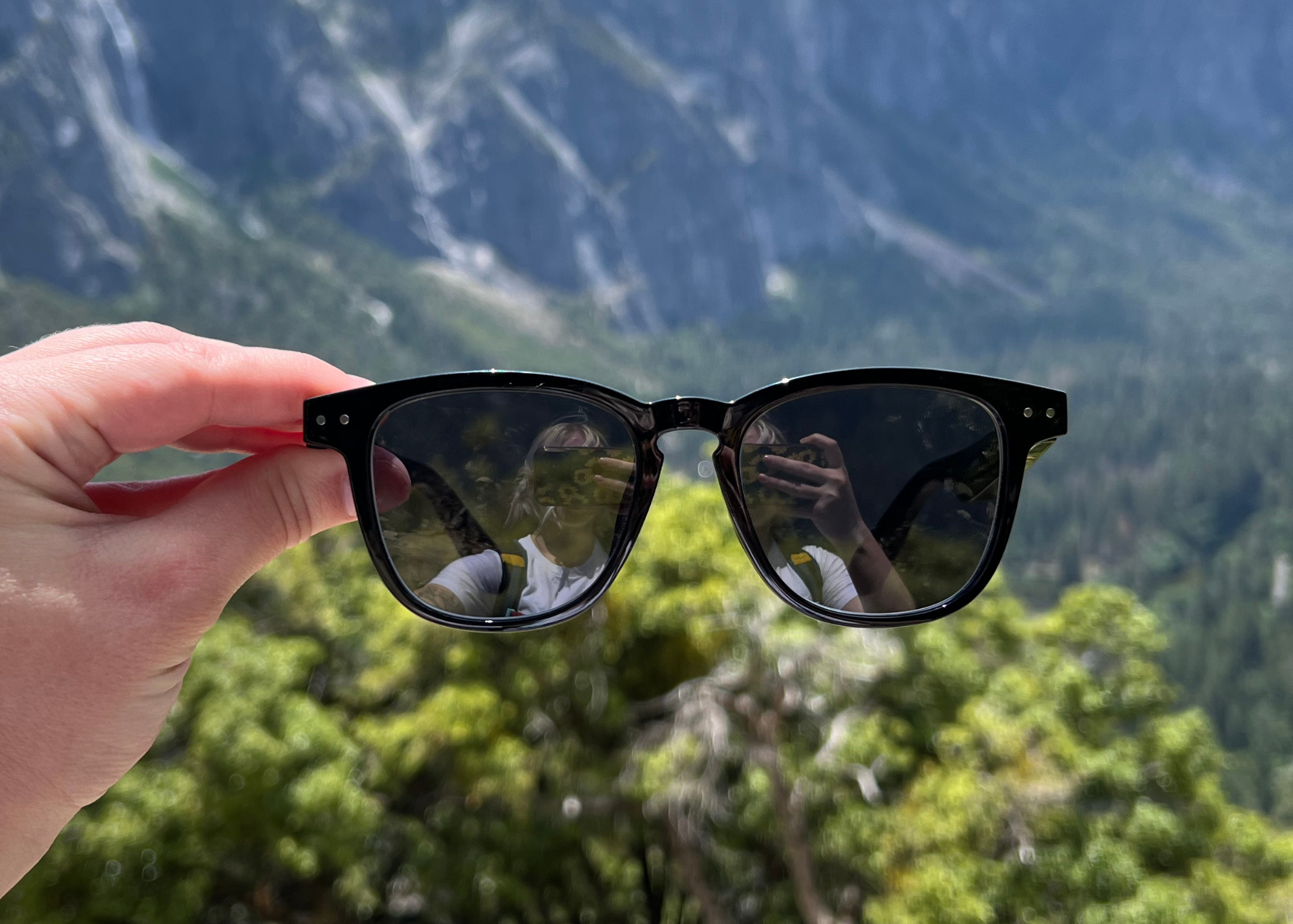 hand holding pair of black sunglasses against mountain background