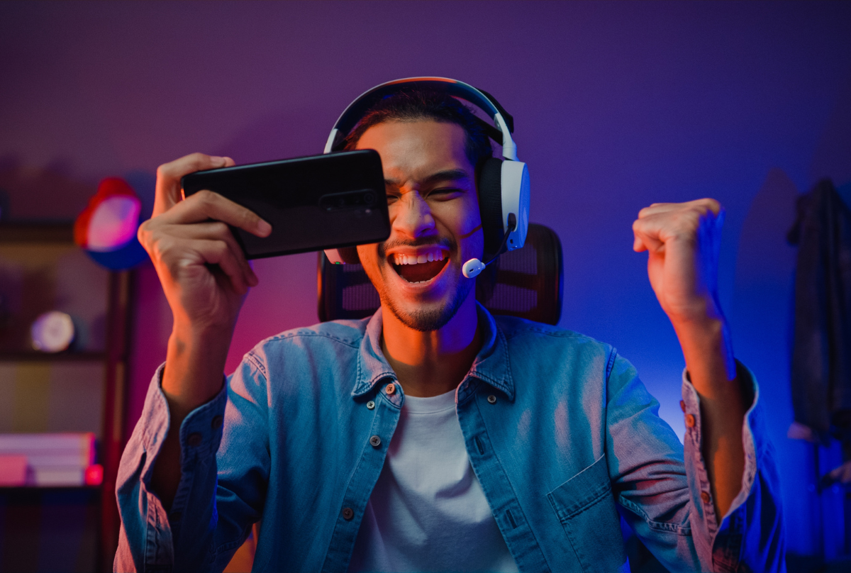 Happy asia man gamer wear headphone competition play video game online with smartphone colorful neon light in living room at night modern house. Esport streaming game online, Home quarantine activity.