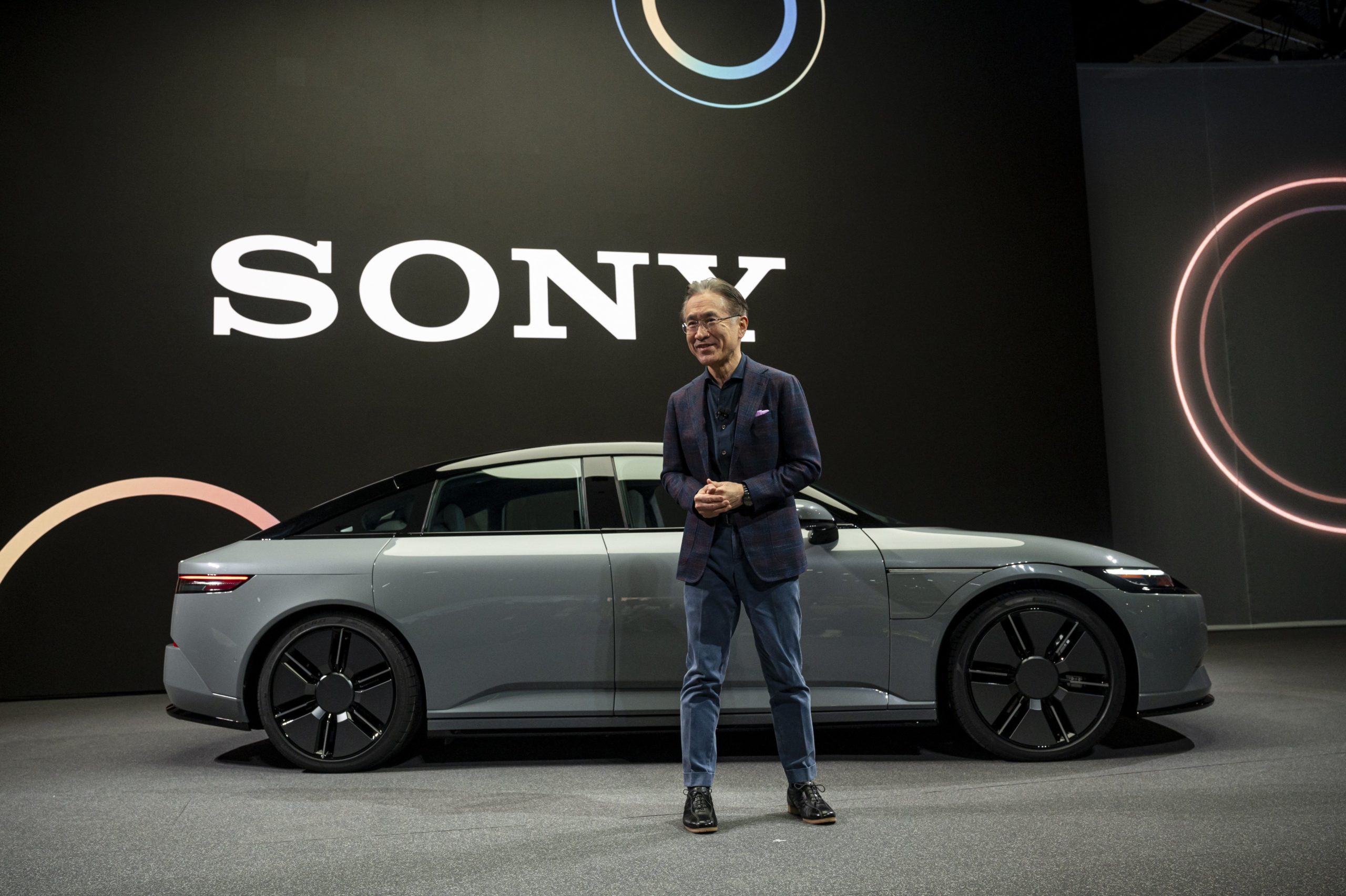 An asian man in a suit stands in front of a car. In the background is a giant screen with the words Sony displayed