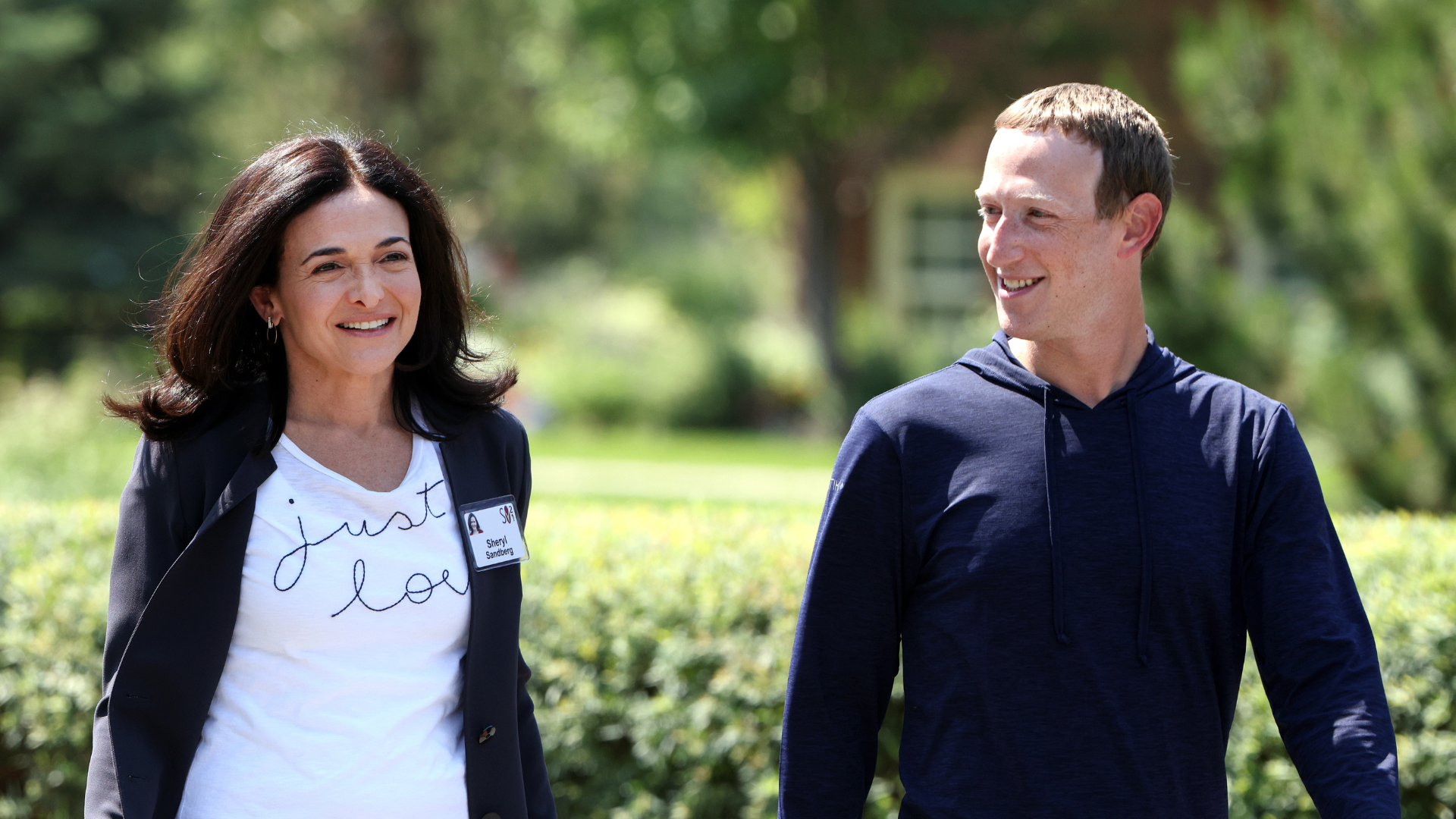 Sheryl Sandberg in a white graphic tee and navy blazer smiles and walks beside Zuckerberg in a navy pullover.