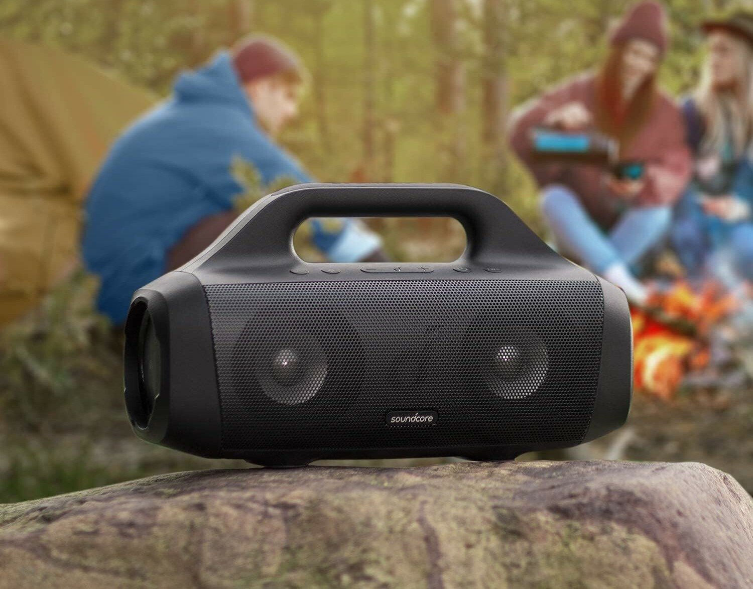 a soundcore anker motion boom speaker sits on a rock outside with people sitting behind it