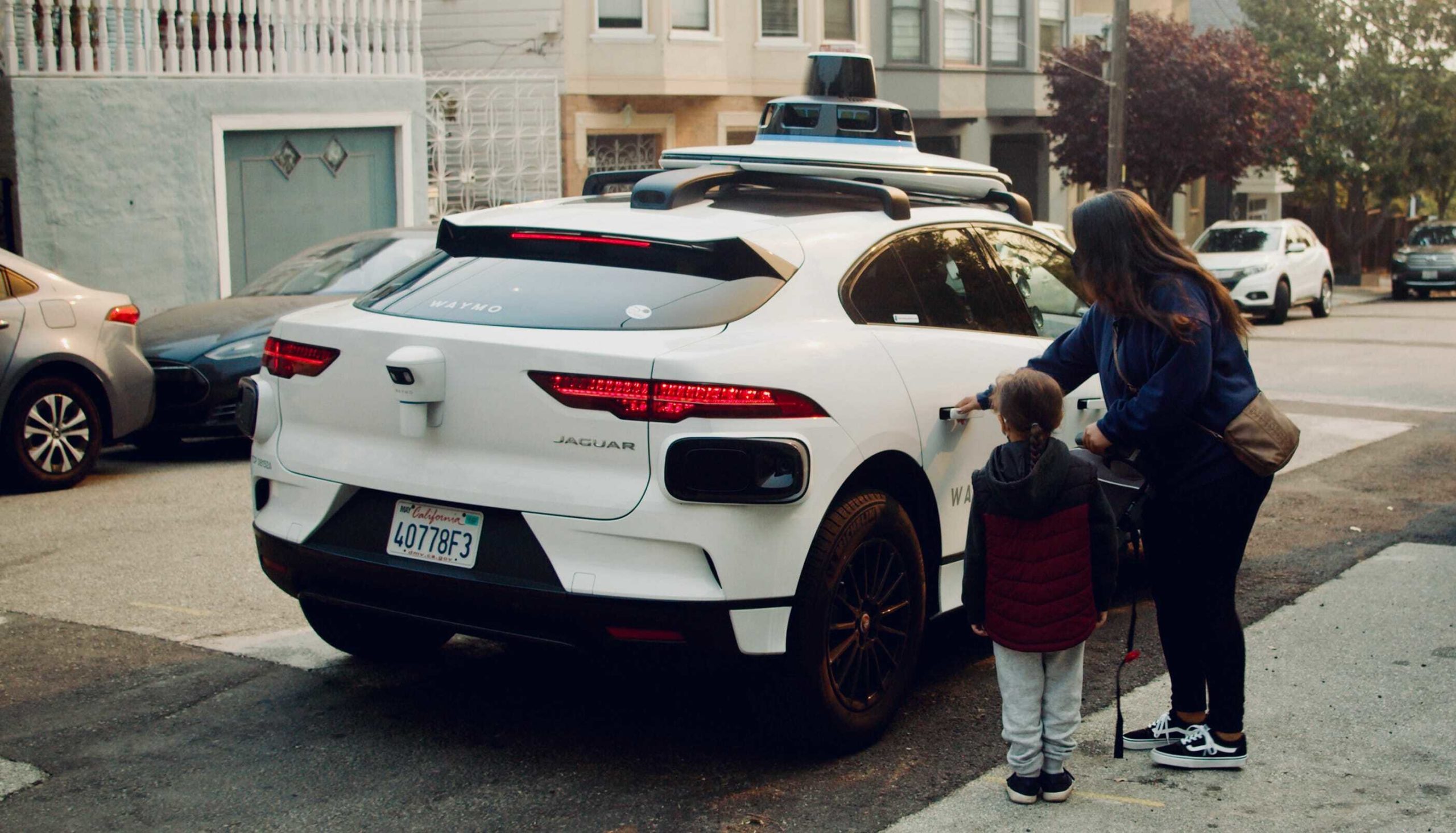 A woman and her child stand outside a white Waymo driverless vehicle, seen from the back. The woman's hand is on the car door's handle and it appears she is about to open the door.