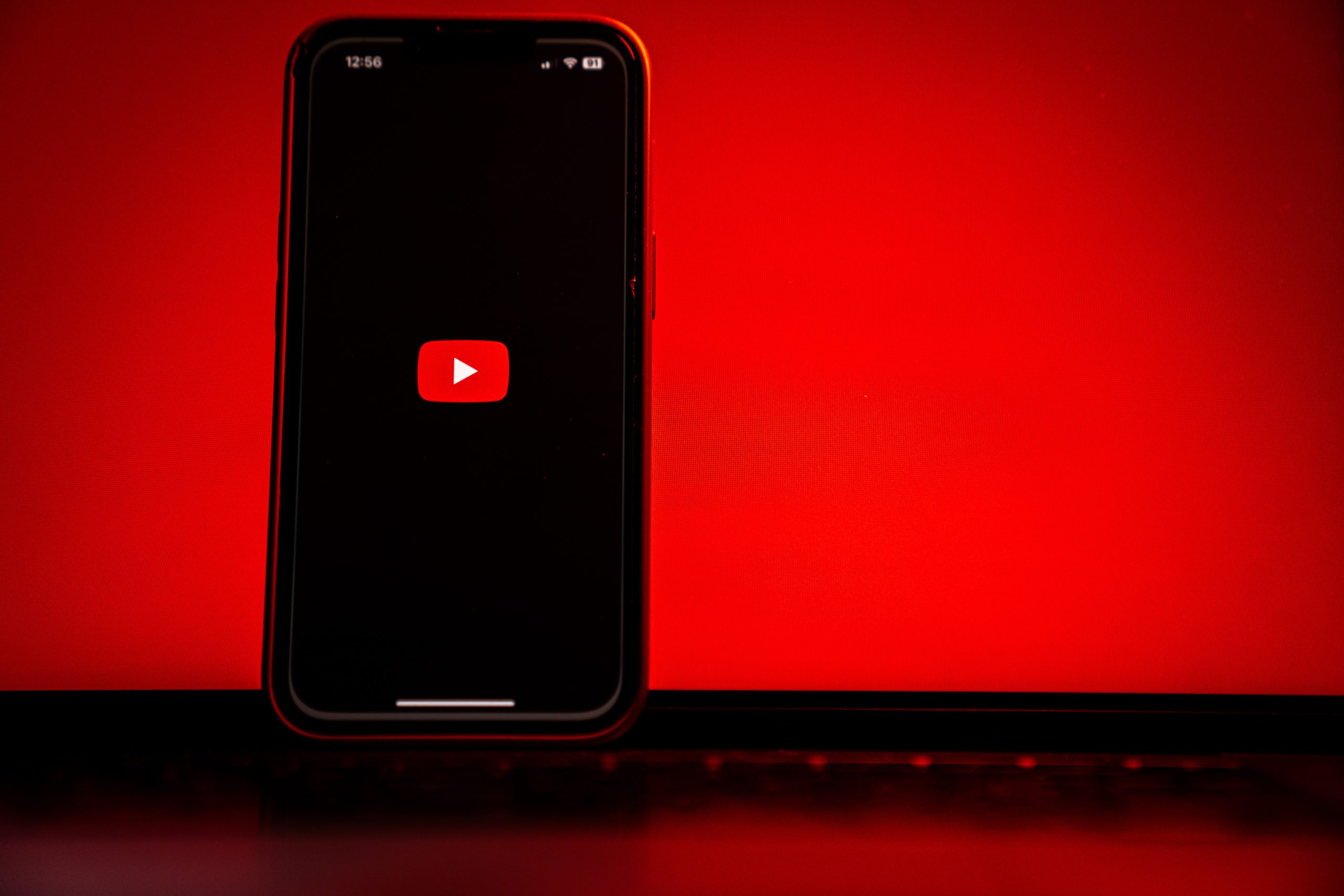 A phone showing the YouTube logo stands propped in front of a glowing red laptop.