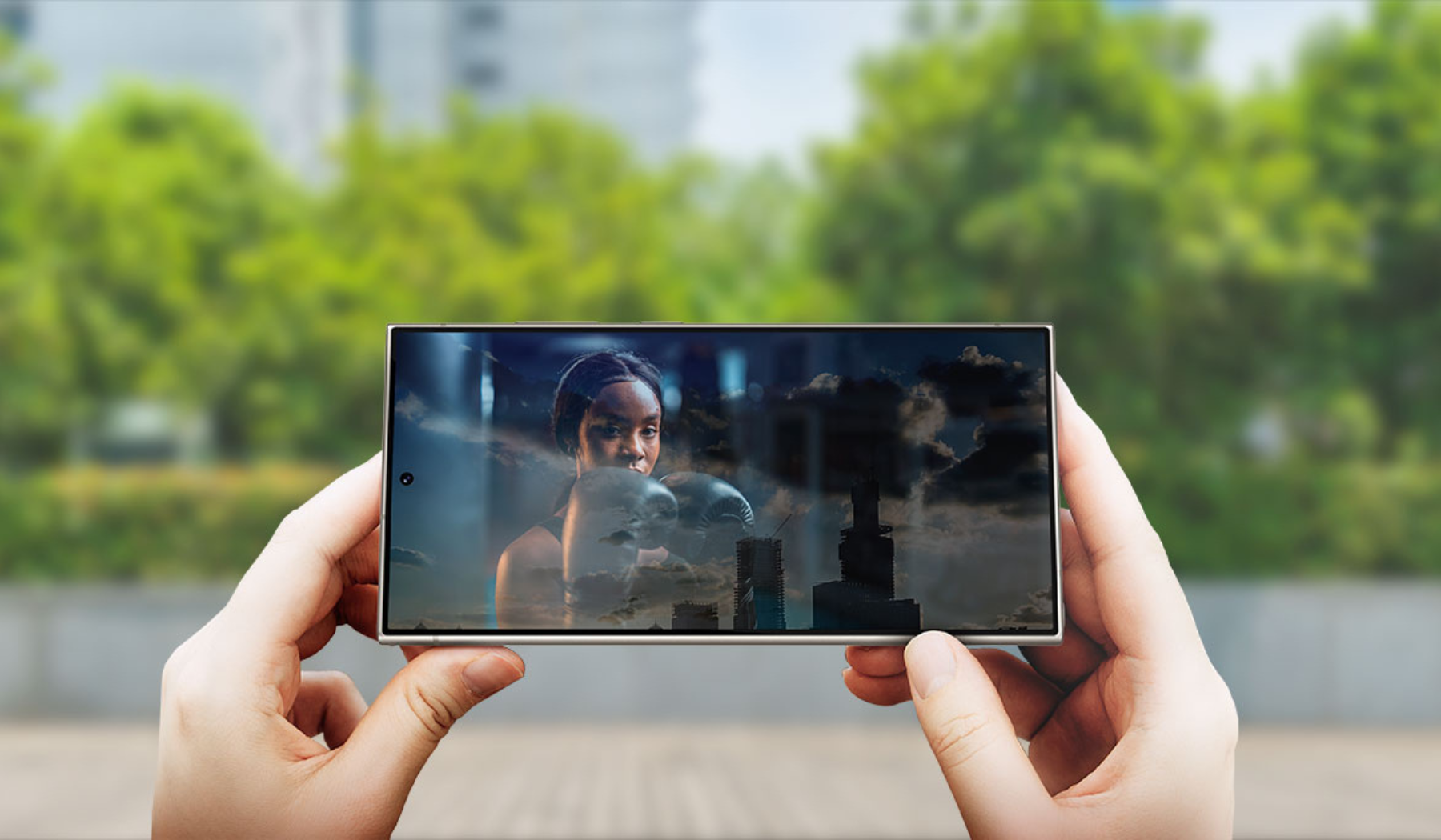 Two hands holding Samsung Galaxy S24 Ultra smartphone with streaming content on screen and trees in background