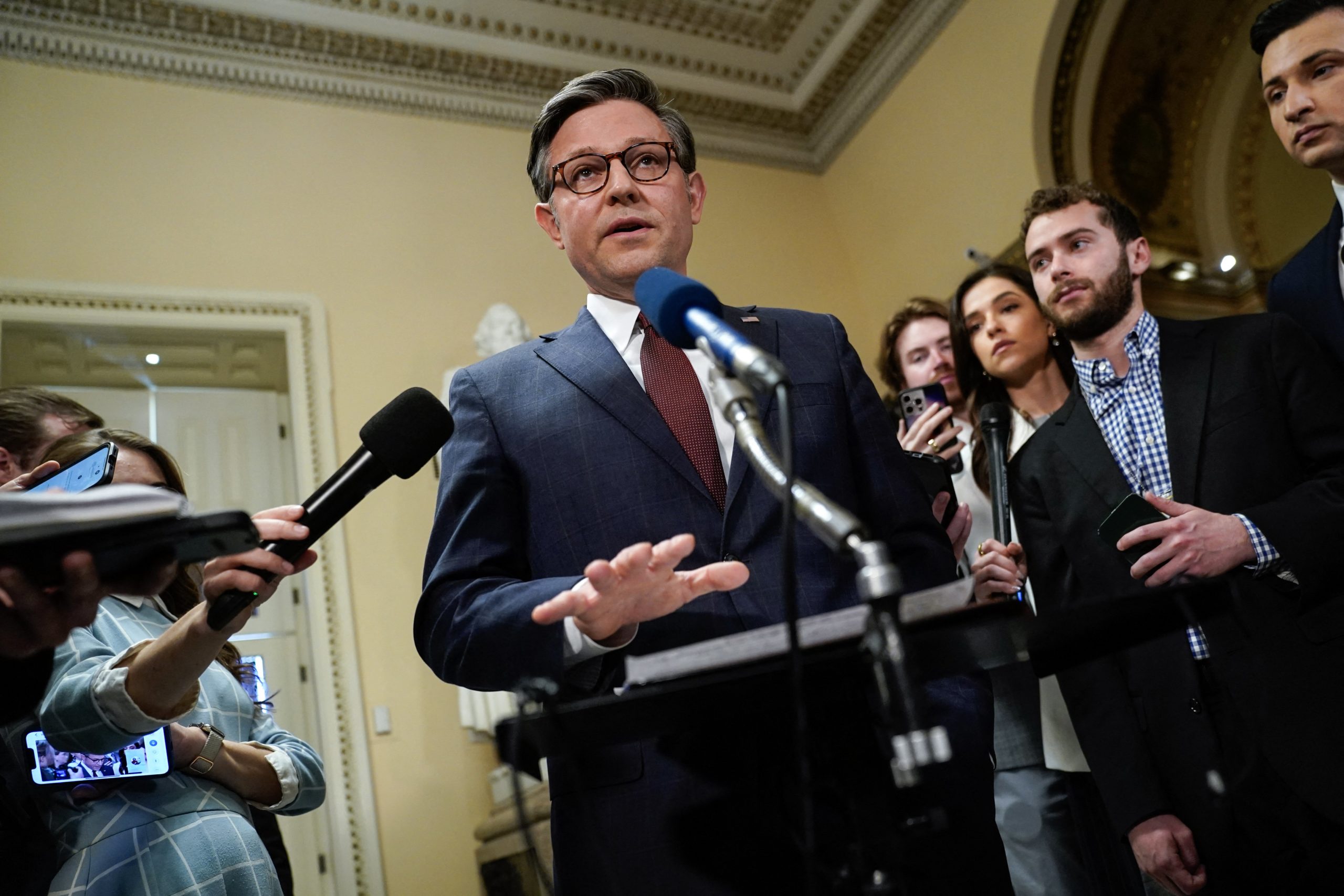 Johnson wears a dark blue suit, white shirt, maroon tie, and spectacles. He stands at a podium/ microphone.