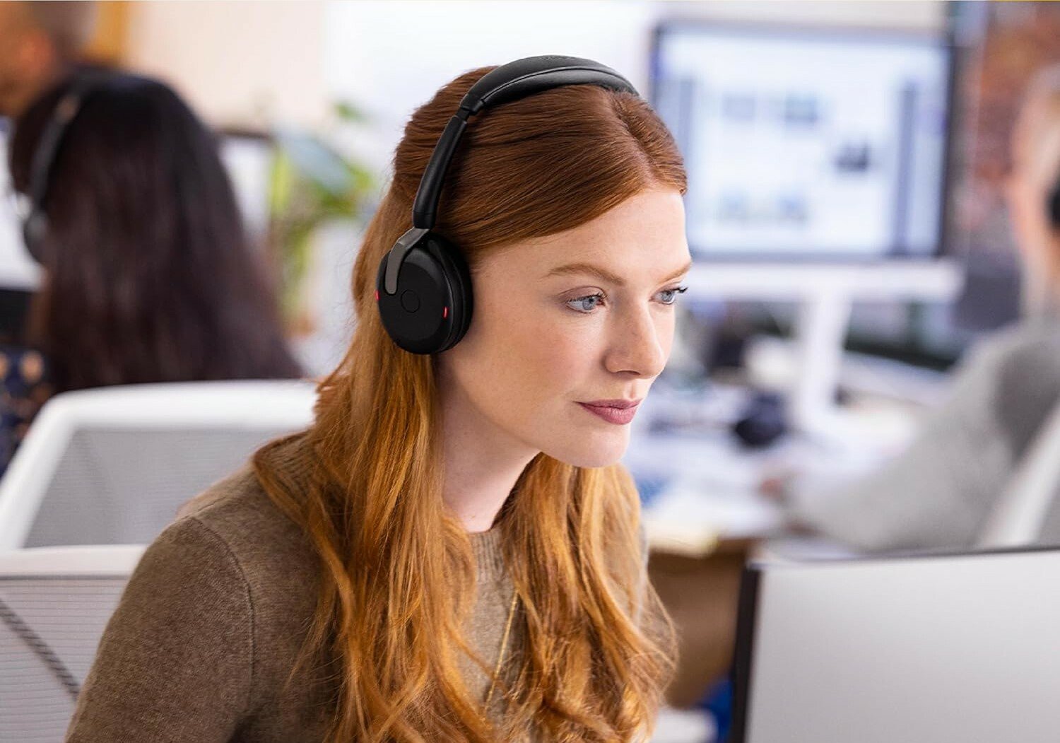 a person with red hair wears the Jabra Evolve2 65 headset while looking at a computer screen. The setting it an office environment with other people working in the background at computer stations.