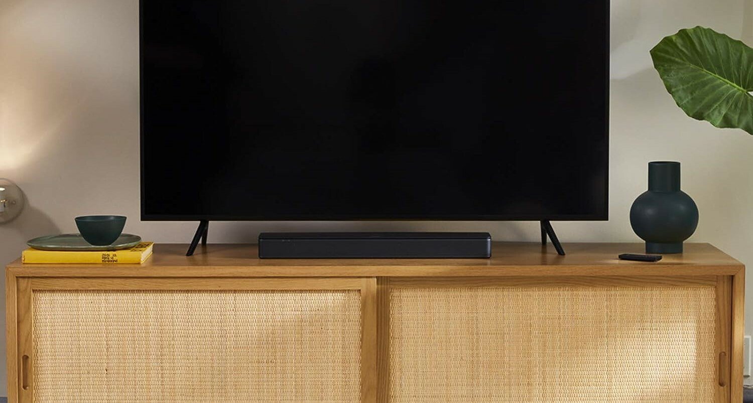 a bose tv speaker soundbar sits below a large TV screen, on top of a light brown entertainment center. The room also has a green plant and the entertainment center has a vase and a stack of books on either side of the TV