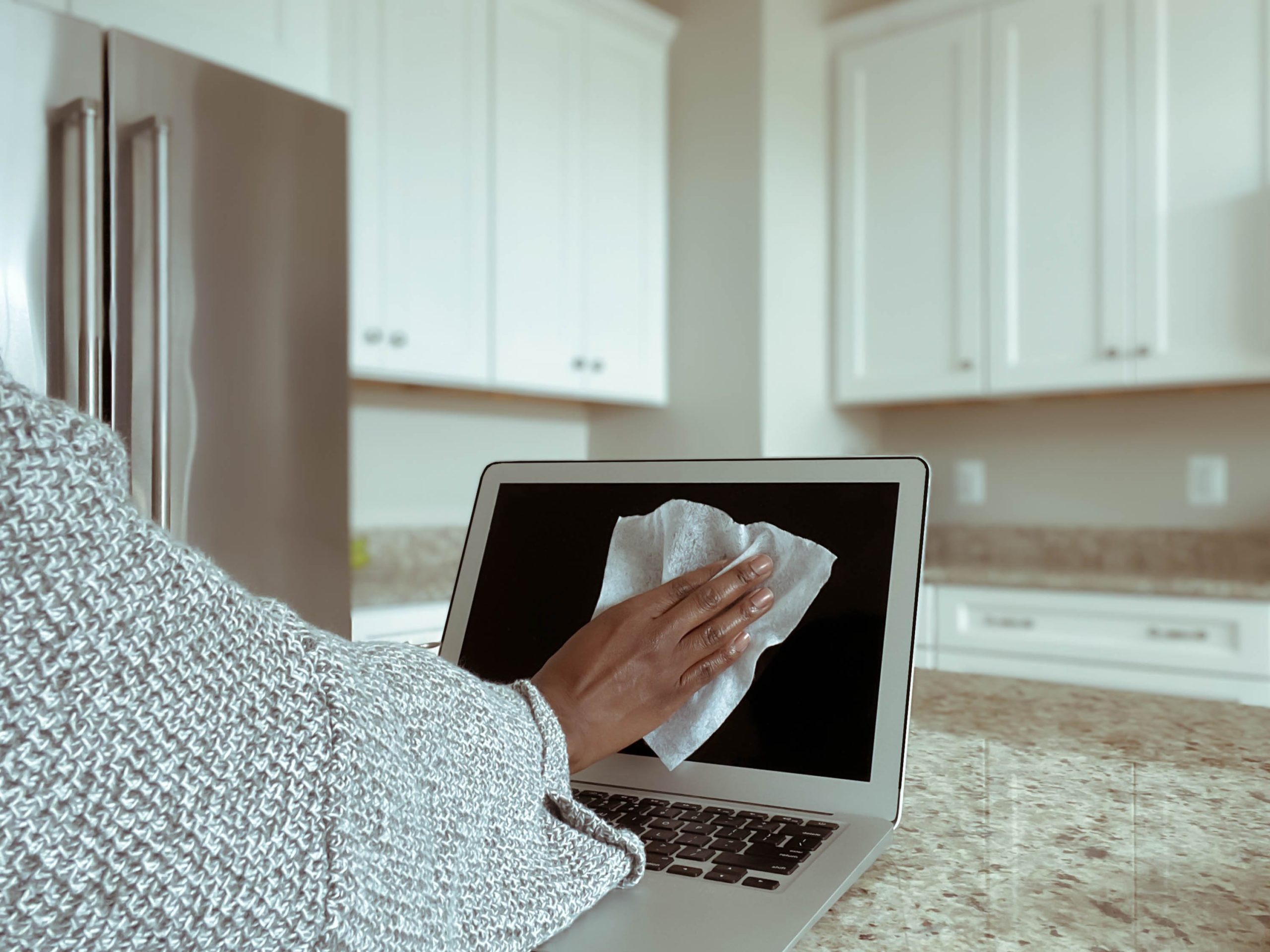 Close-up of unrecognizable woman in kitchen cleaning laptop screen with disinfectant wipe