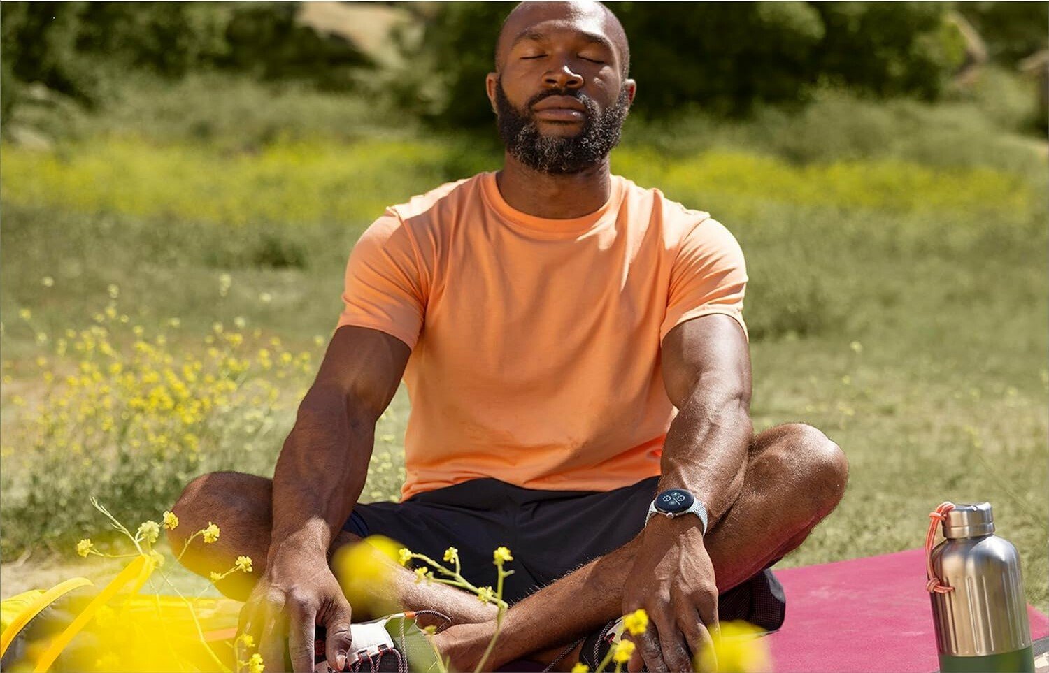 a person wearing an orange shirt and black shorts sits cross-legged in a field of grass while wearing the google pixel 2 watch on his left wrist.