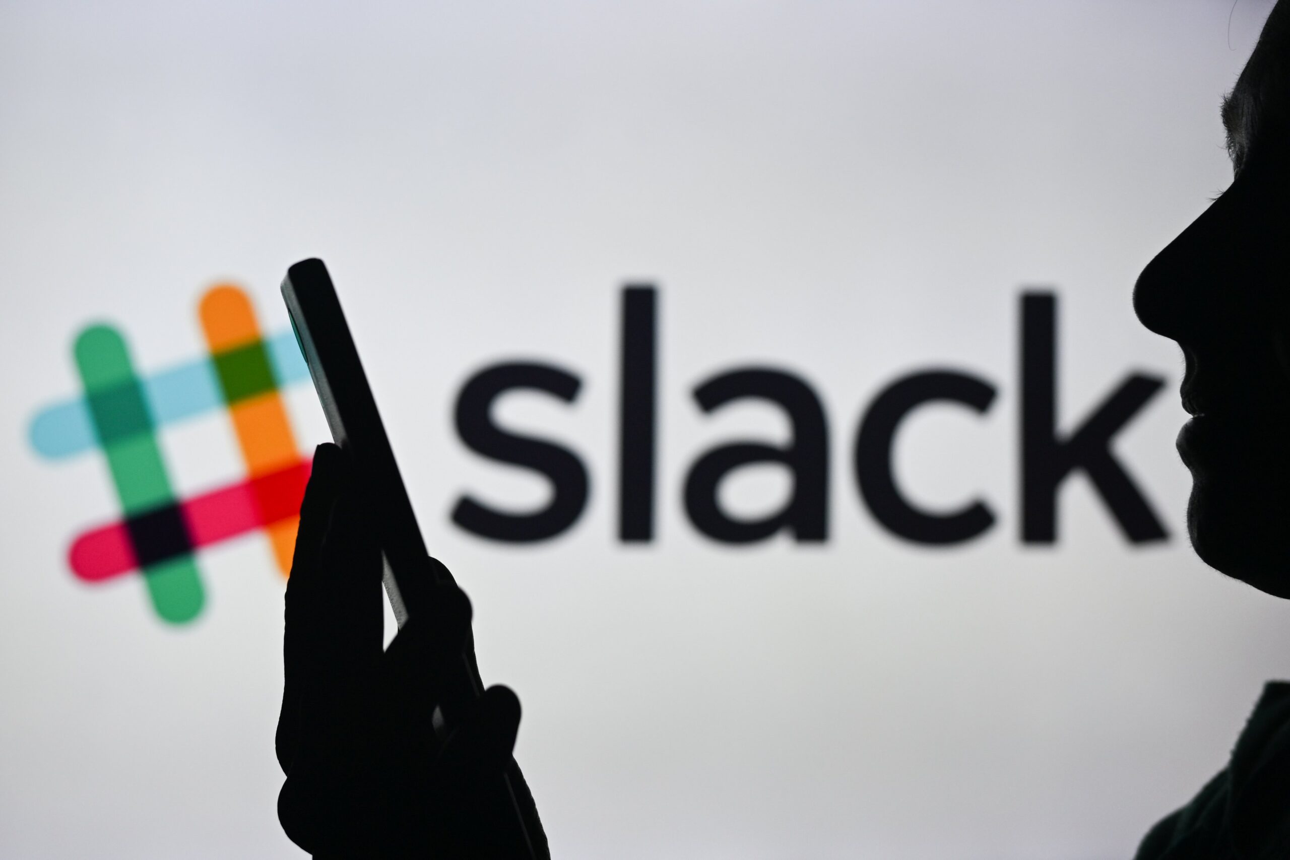 A woman holding a cell phone in front of the Slack logo displayed on a computer screen.