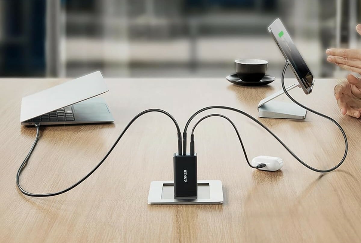 an anker charger with three ports sits on a desk while charging a laptop, cell phone, and earbuds