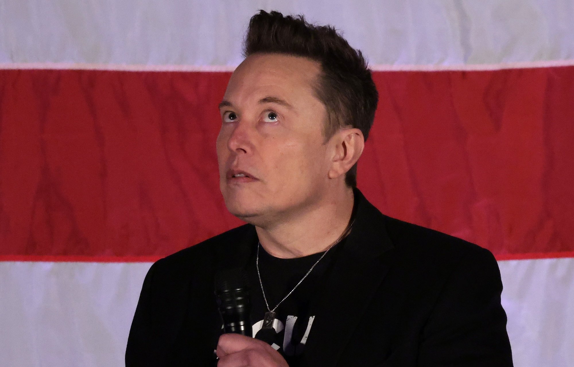 Elon Musk stares upwards in front of a US flag. 