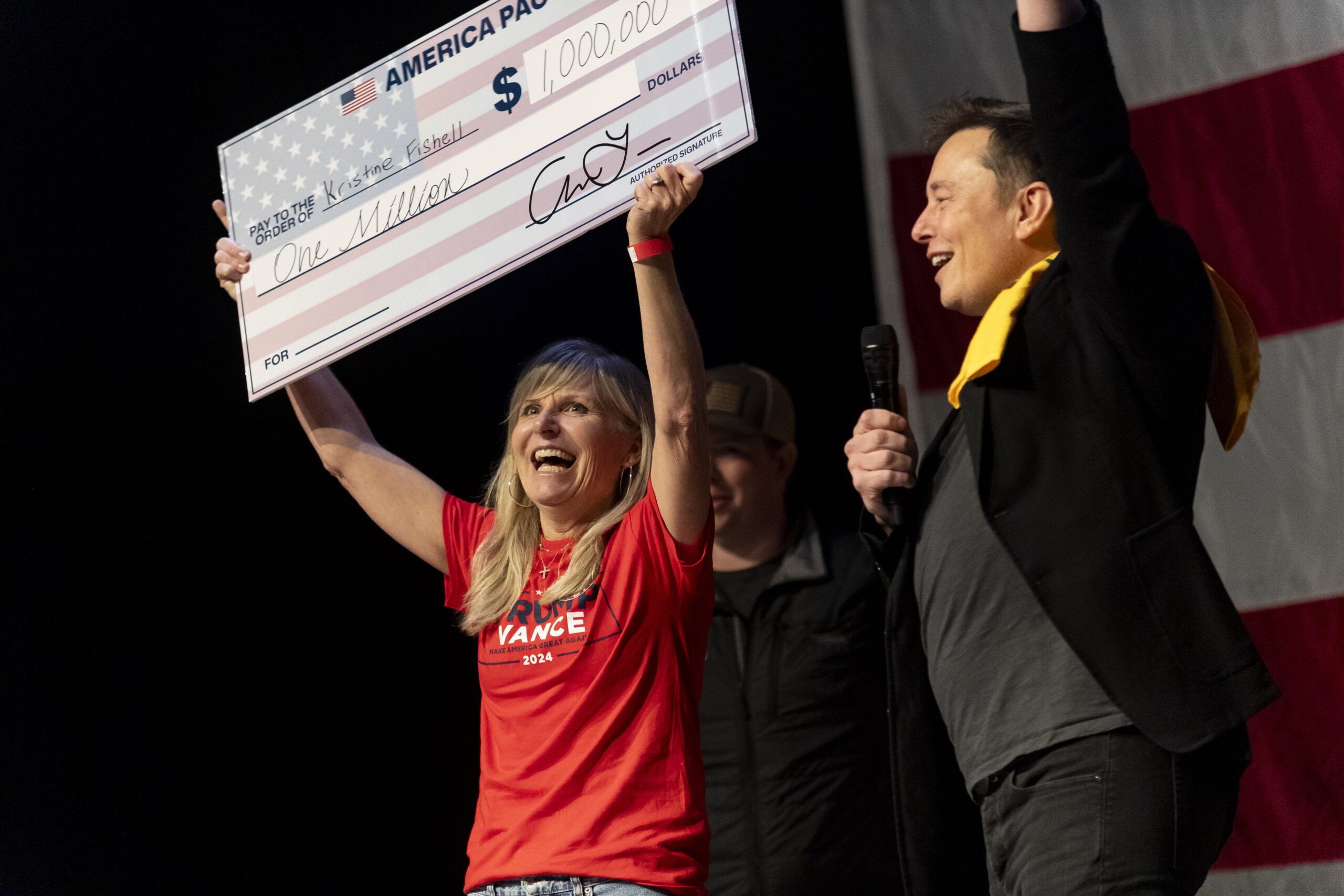 Elon Musk stands on stage cheering for a woman holding up a giant check written out for $1 million.