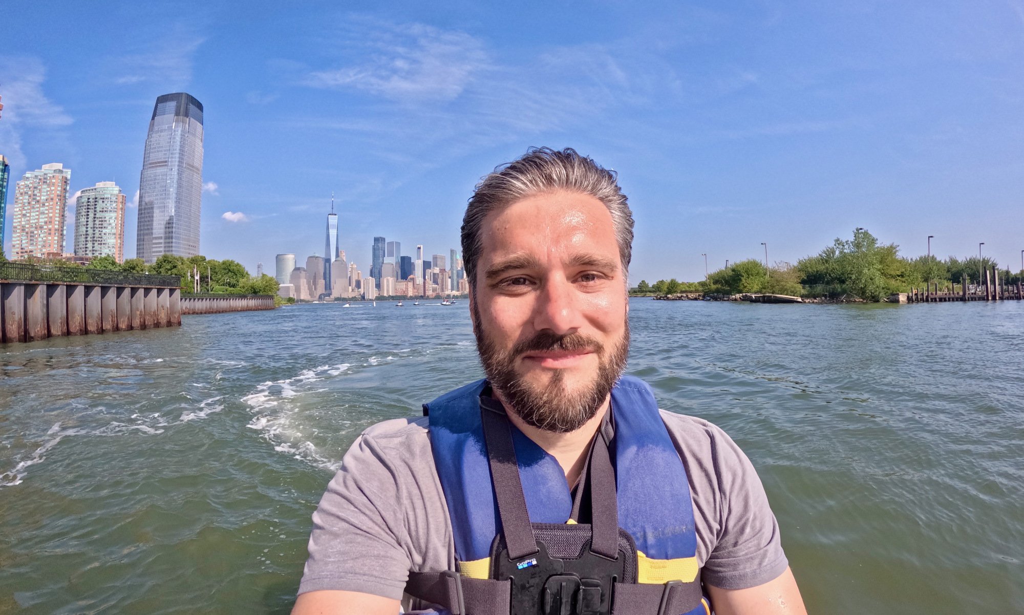 a very handsome man with windswept salt-and-pepper hair smiles while on a jet ski with new york city skyline in background