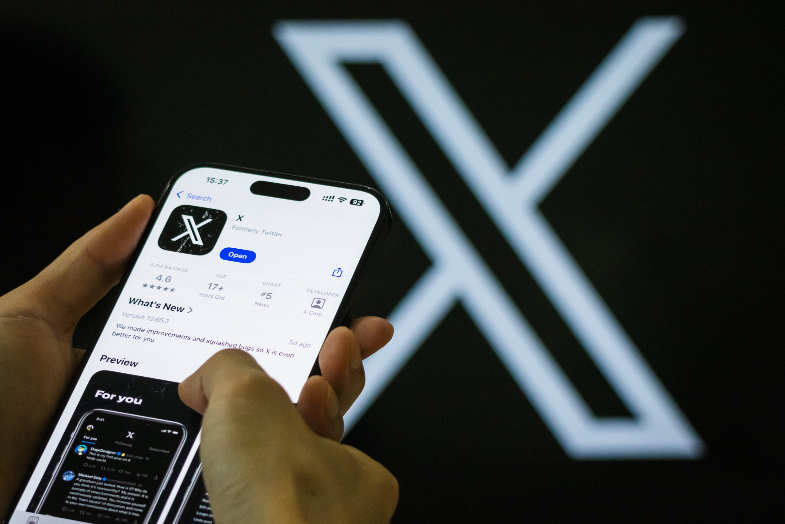 A user interacts with the X app on a smartphone, showcasing the app’s rebranded interface, in front of the X logo