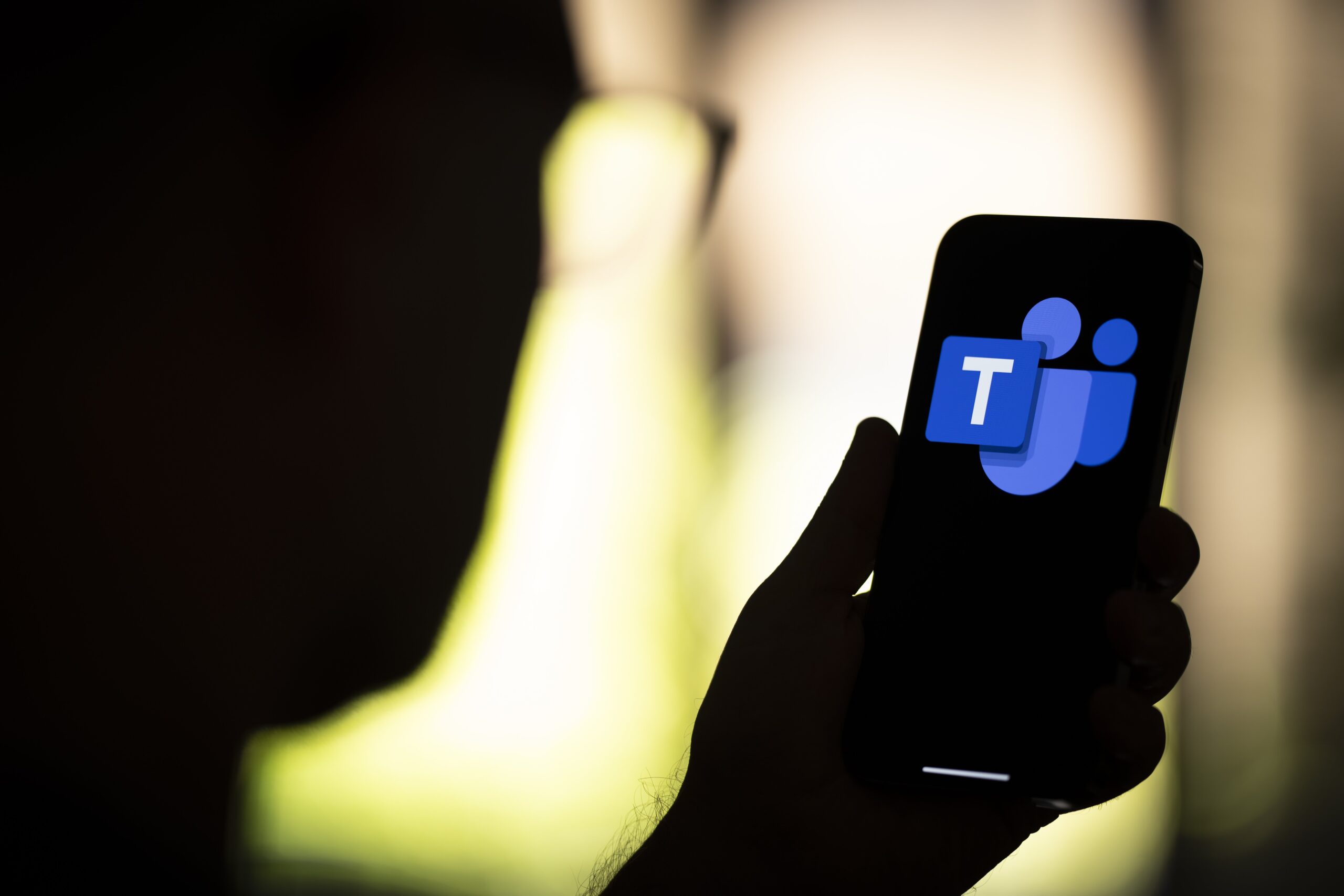 A person in silhouette holds up a phone displaying the blue Microsoft Teams logo.