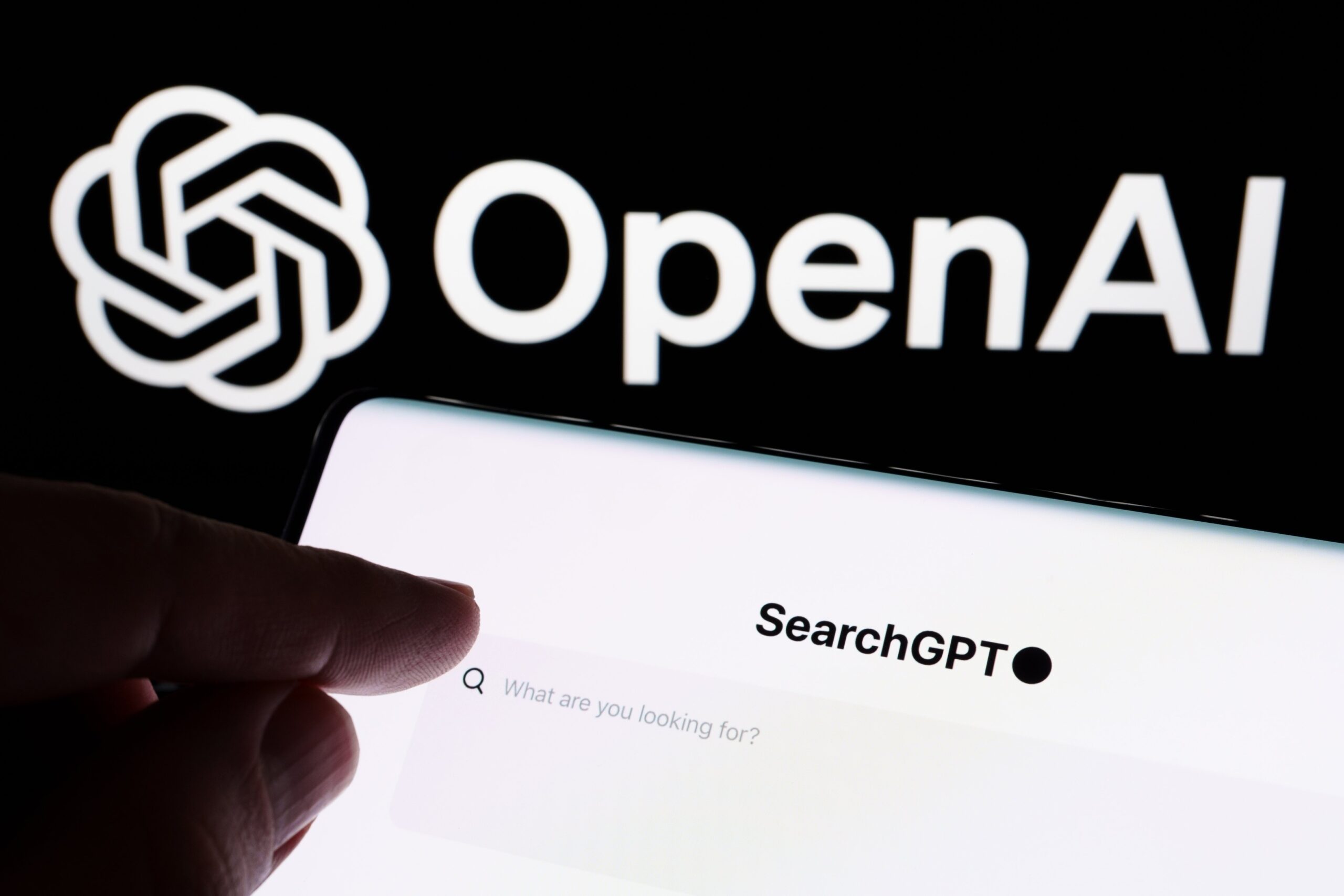This image shows a close-up of a smartphone screen displaying the text “SearchGPT”; In the background, the OpenAI logo is prominently displayed,