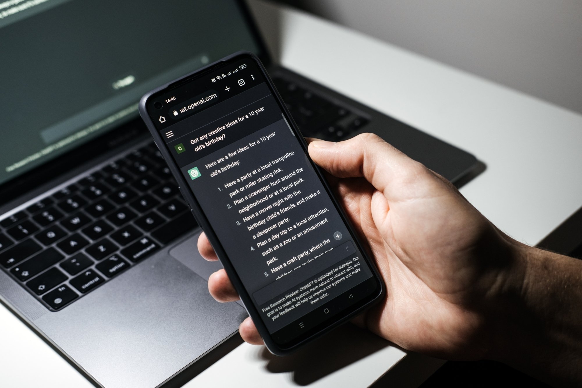 Person using ChatGPT on a phone with a laptop in the background