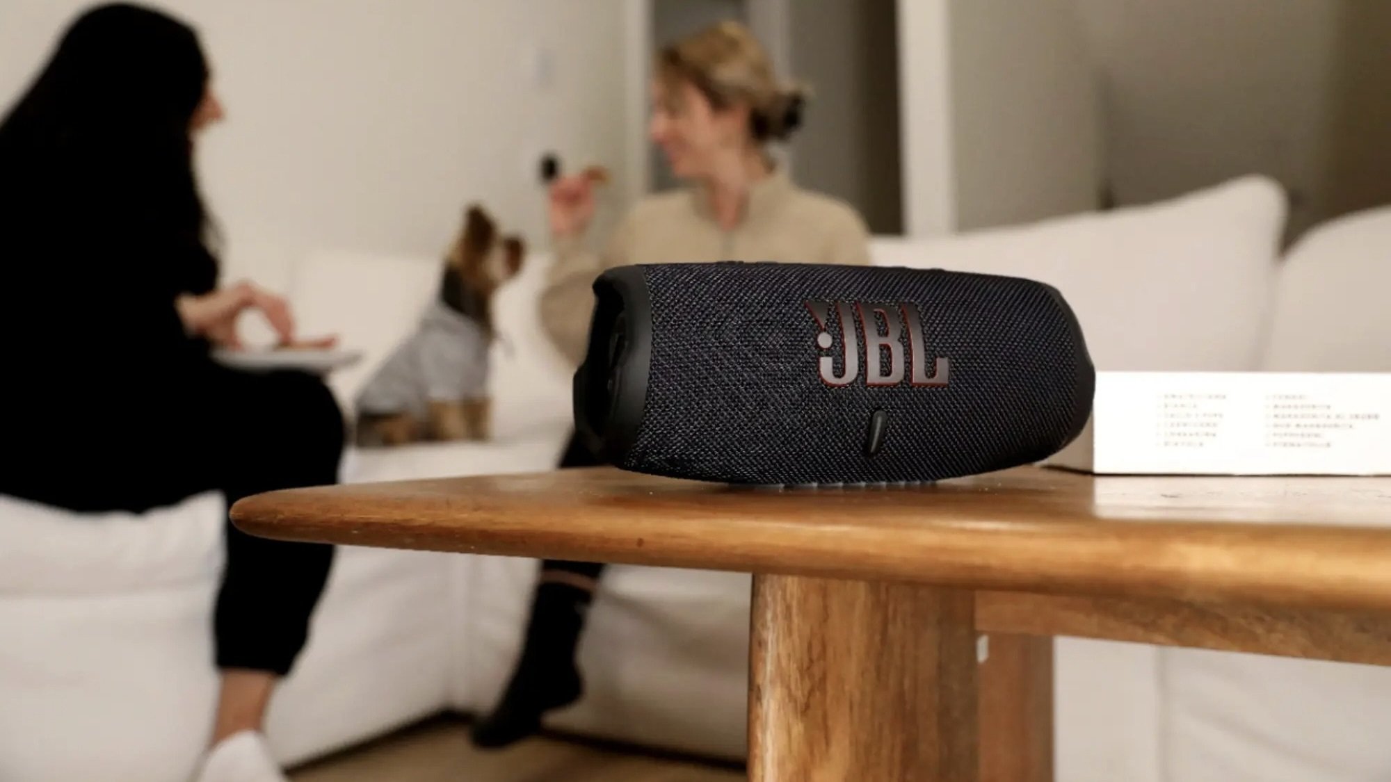 jbl charge 5 speaker on coffee table in front of two women and dog sitting on living room couch