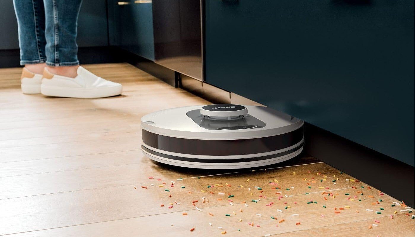 the shark ai robot vacuum cleans colorful sprinkles off a kitchen floor while someone wearing white slip-on shoes stands in the background