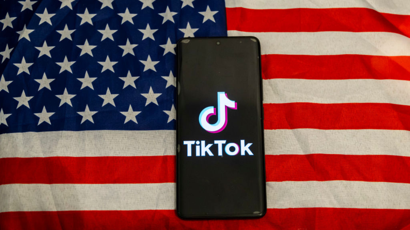 The TikTok app logo on a smartphone screen. The phone is laying on an American flag.